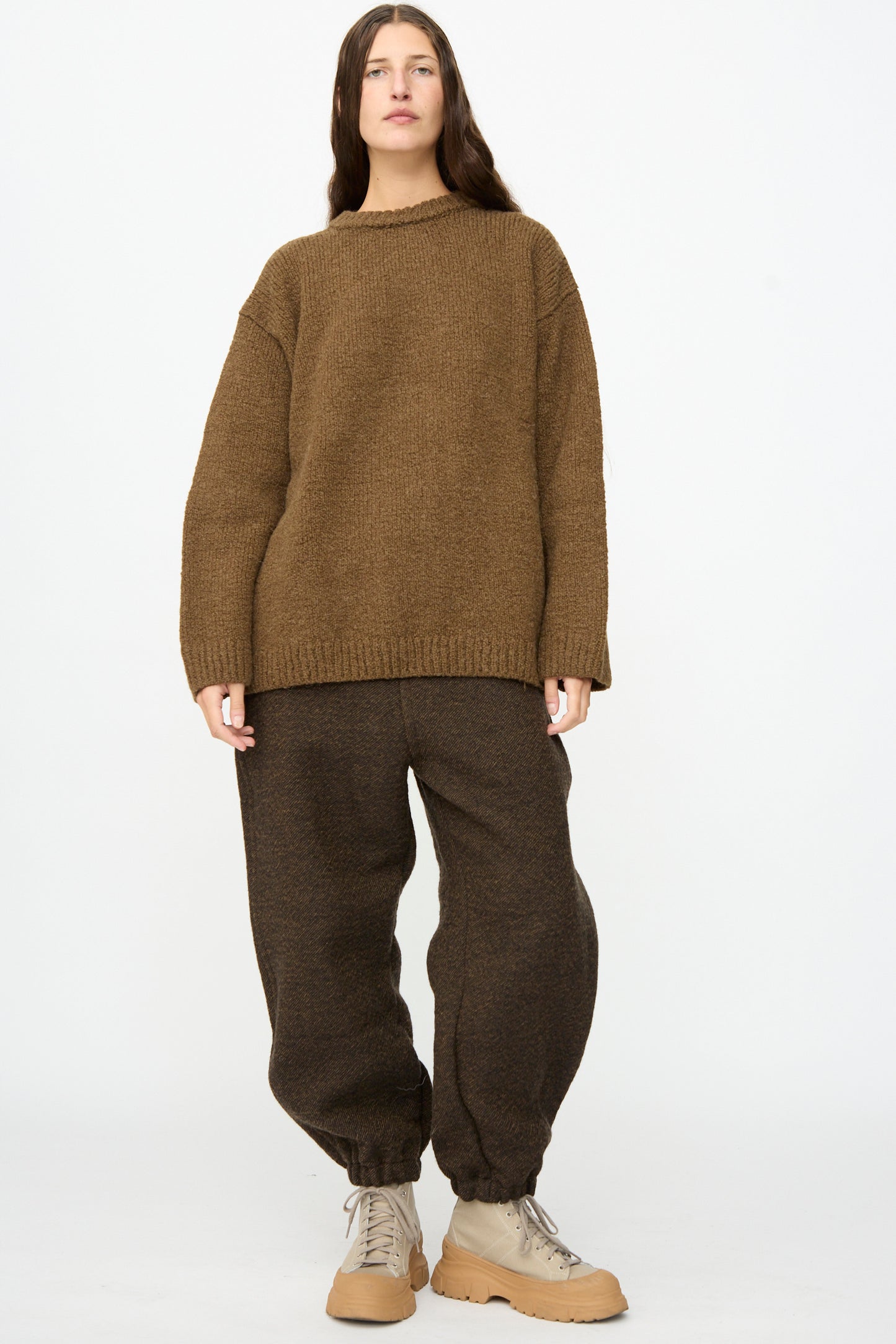 A person stands against a plain background, wearing the SONO Jako Jumper in Kelp—an oversized pullover sweater crafted from a luxurious wool alpaca blend—paired with dark brown baggy pants featuring elastic cuffs and beige lace-up boots.
