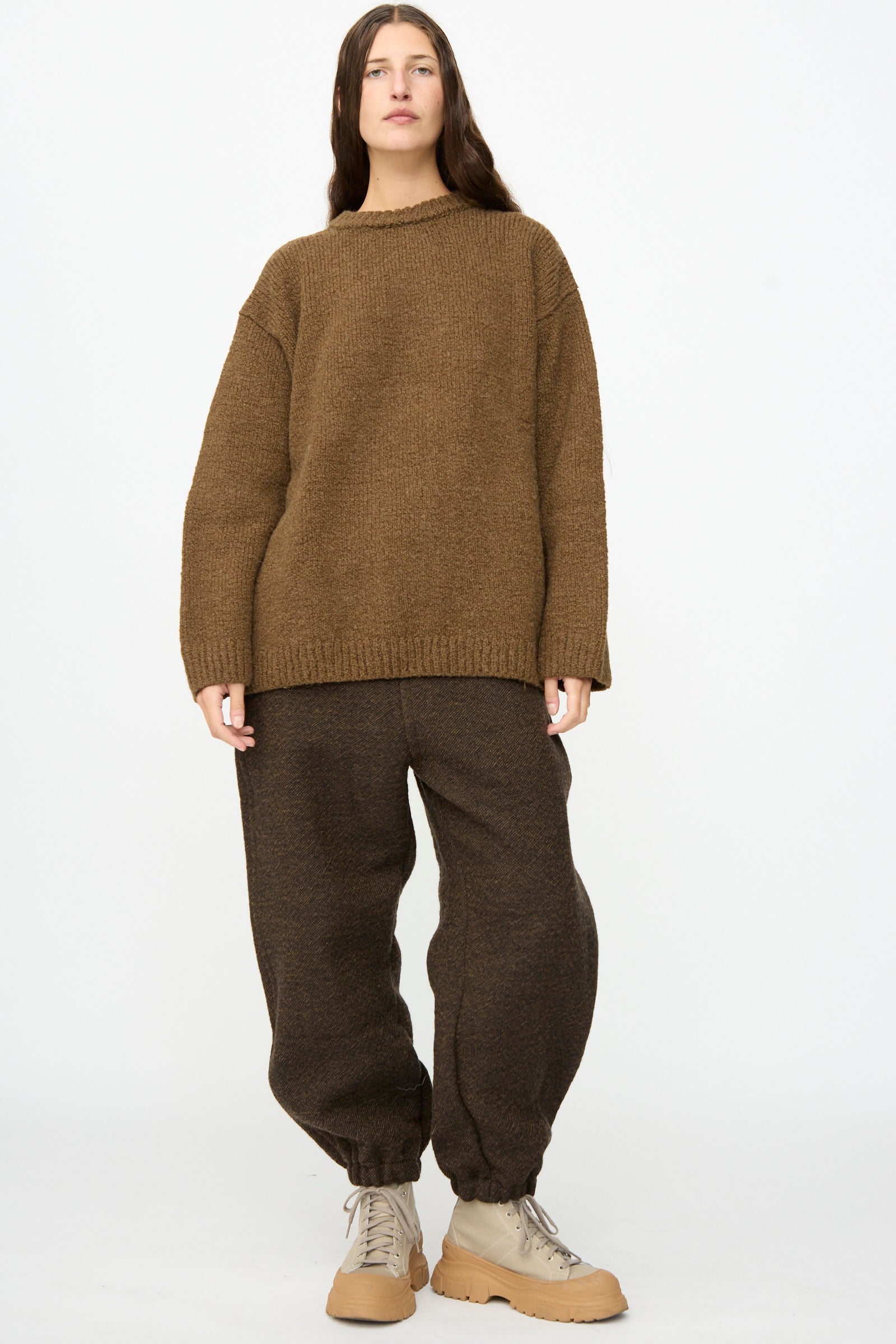 A person stands against a plain background, wearing the SONO Jako Jumper in Kelp—an oversized pullover sweater crafted from a luxurious wool alpaca blend—paired with dark brown baggy pants featuring elastic cuffs and beige lace-up boots.