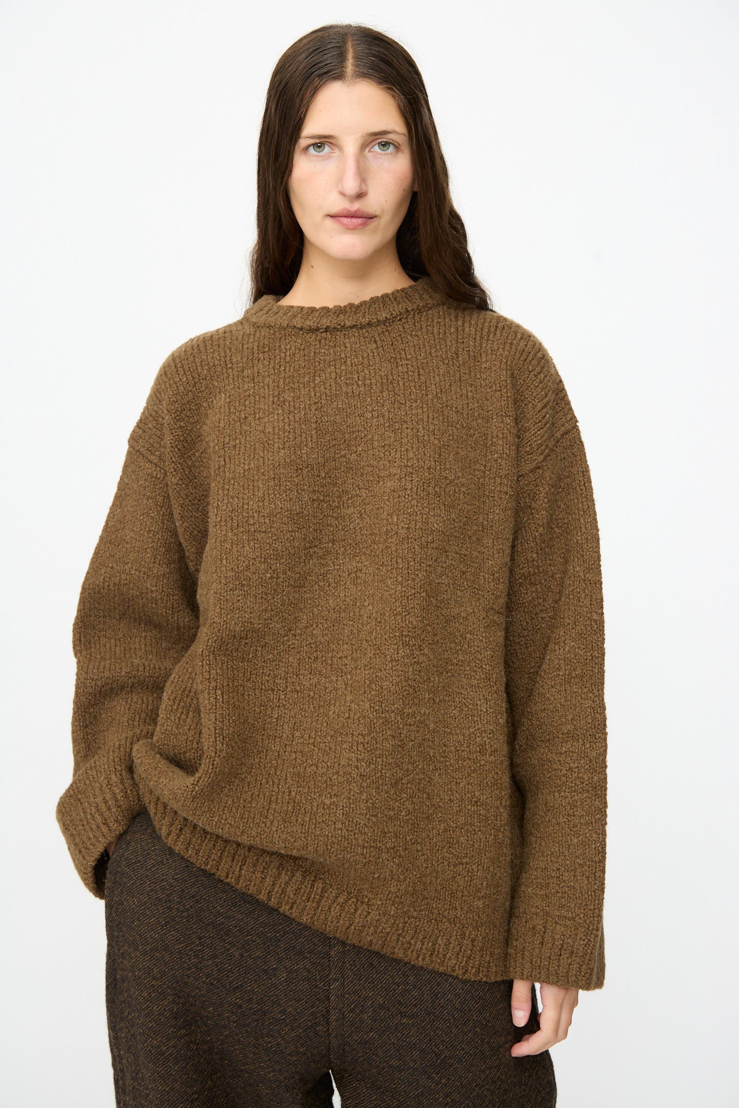 A person with long brown hair wears the SONO Jako Jumper in Kelp—an oversized pullover sweater crafted from a luxurious wool alpaca blend—paired with dark brown pants, standing against a plain white background.