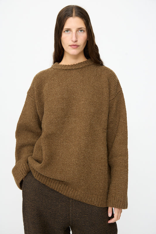 A person with long brown hair wears the SONO Jako Jumper in Kelp—an oversized pullover sweater crafted from a luxurious wool alpaca blend—paired with dark brown pants, standing against a plain white background.