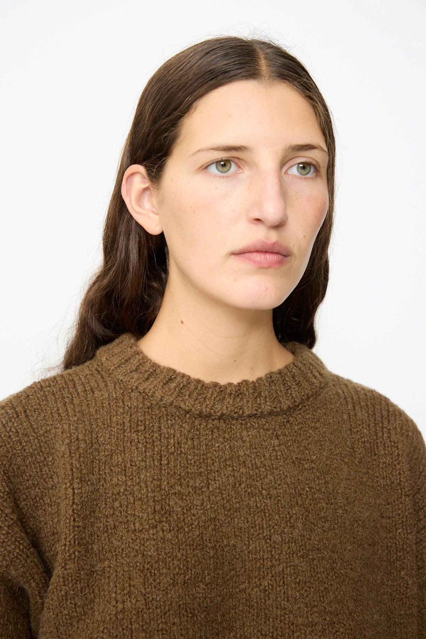 A person with long brown hair and a neutral expression wears the SONO Jako Jumper in Kelp—an oversized pullover sweater crafted from a textured brown wool alpaca blend, made in the UK—against a plain white background.