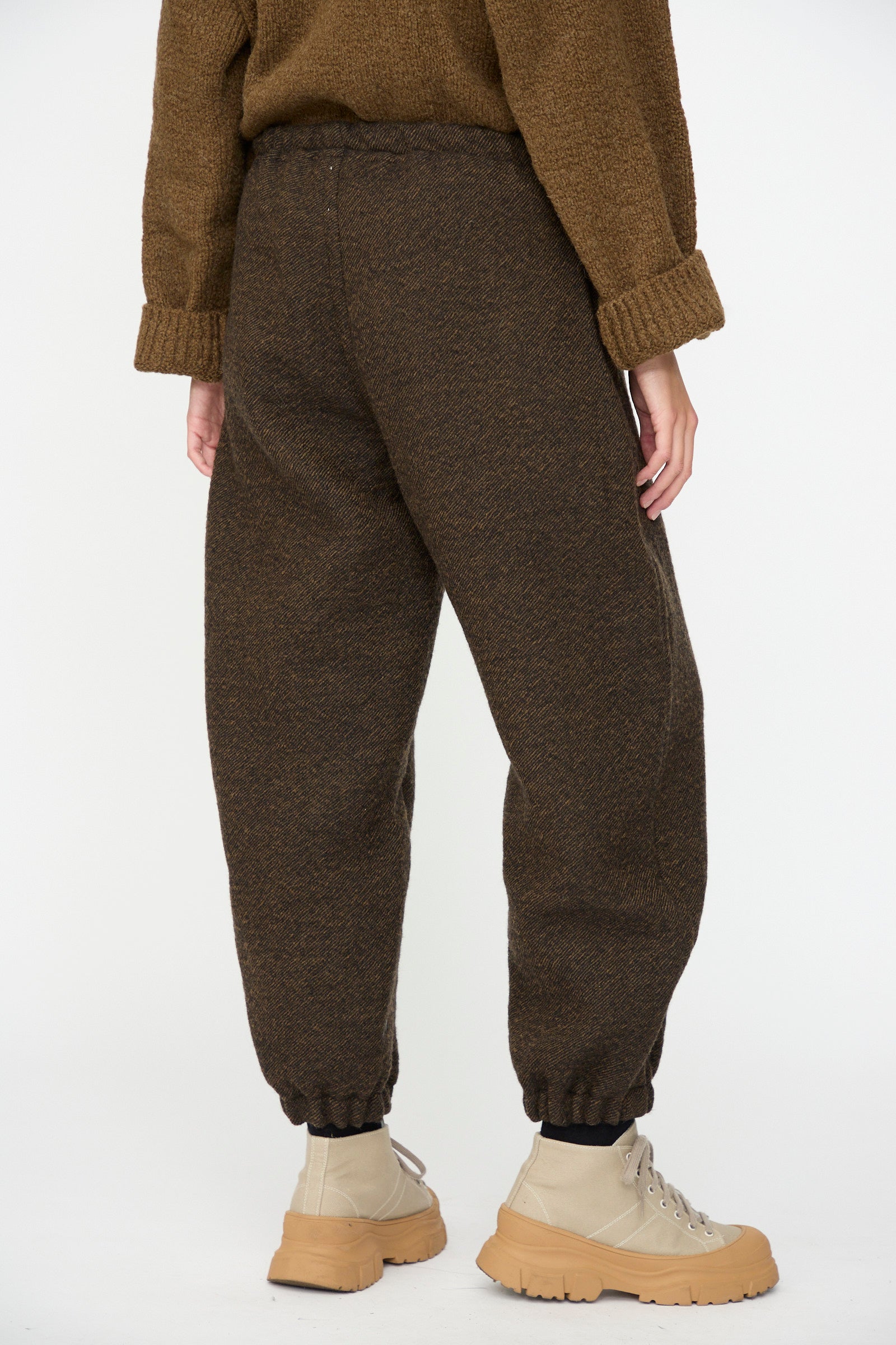 Person wearing SONO’s Jamie Joggers in Blackmud—a pair of brown, textured wool jogger pants—styled with beige lace-up boots and a brown sweater. The photo, taken from behind, showcases the lower half of the body against a plain background. Made in France, these stylish wool joggers offer an effortlessly refined look.