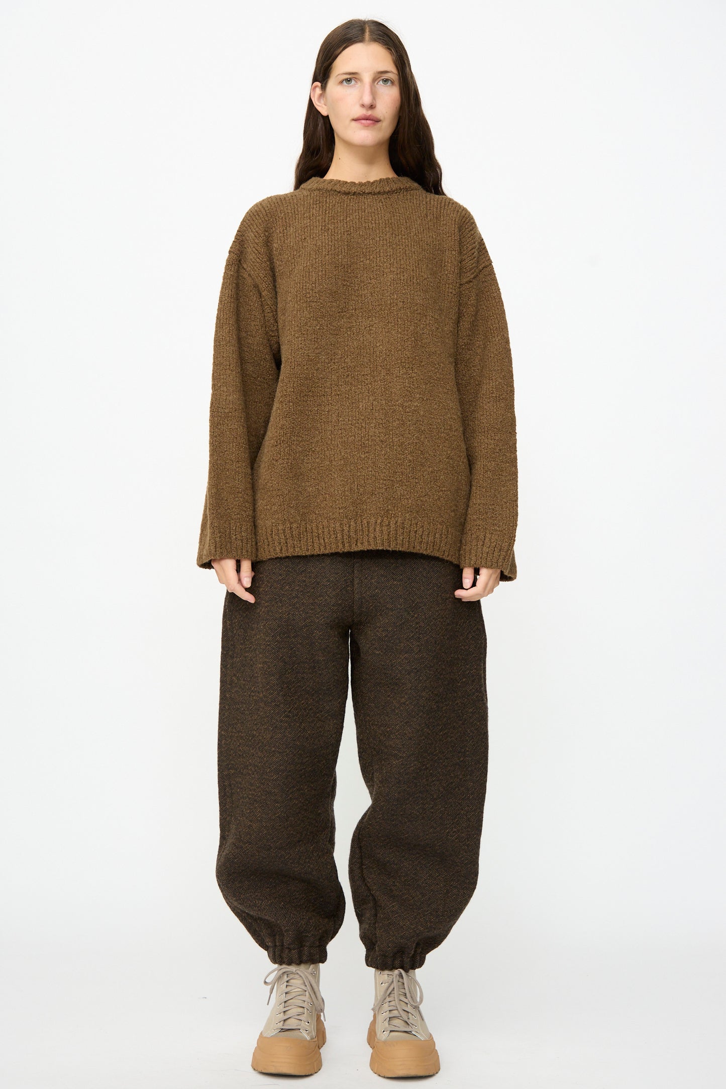 A person stands facing forward, wearing SONO’s loose Jamie Joggers in Blackmud, baggy and cinched at the ankles, paired with a brown sweater and beige lace-up shoes against a plain white background.