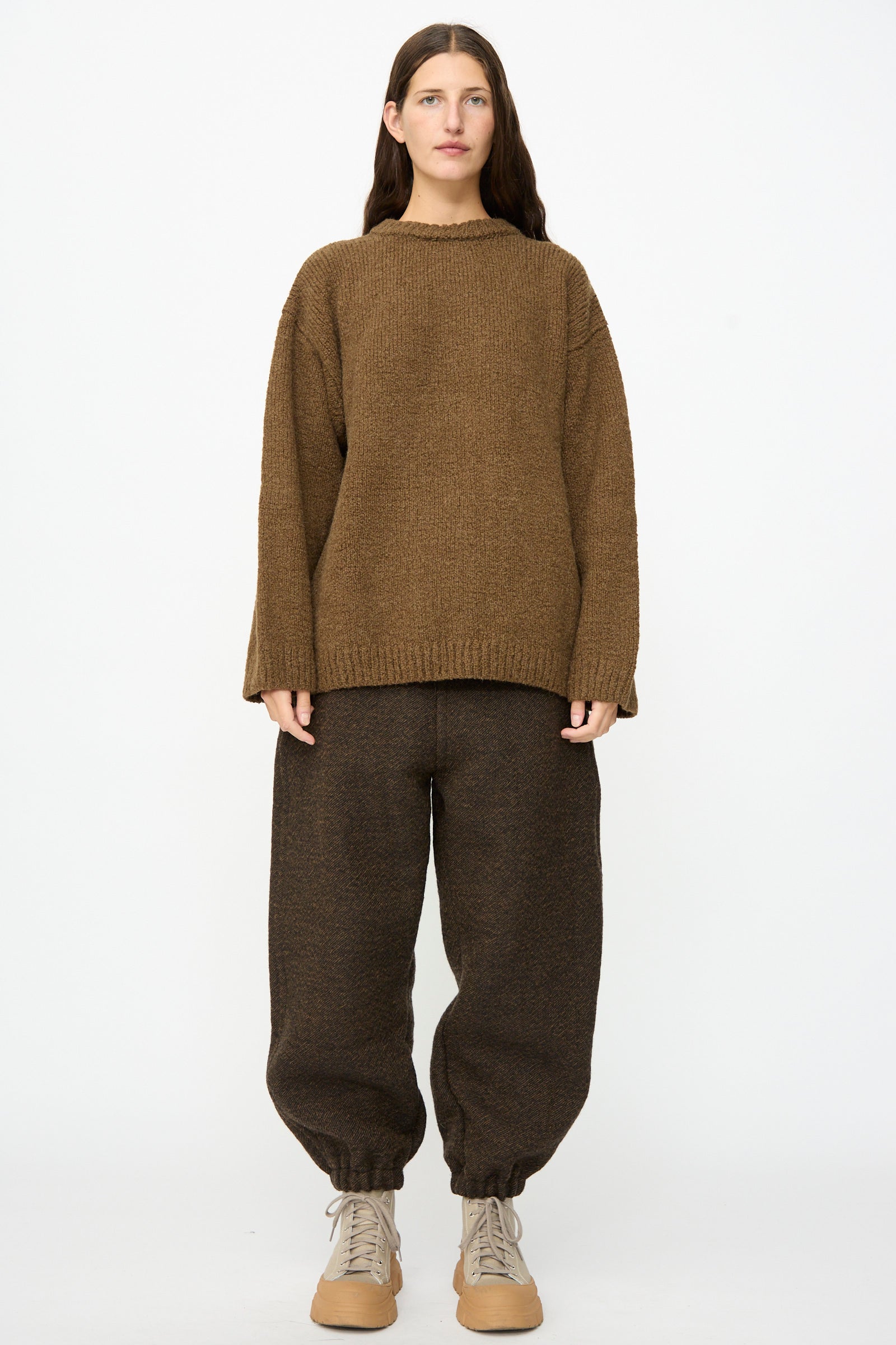 A person stands facing forward, wearing SONO’s loose Jamie Joggers in Blackmud, baggy and cinched at the ankles, paired with a brown sweater and beige lace-up shoes against a plain white background.