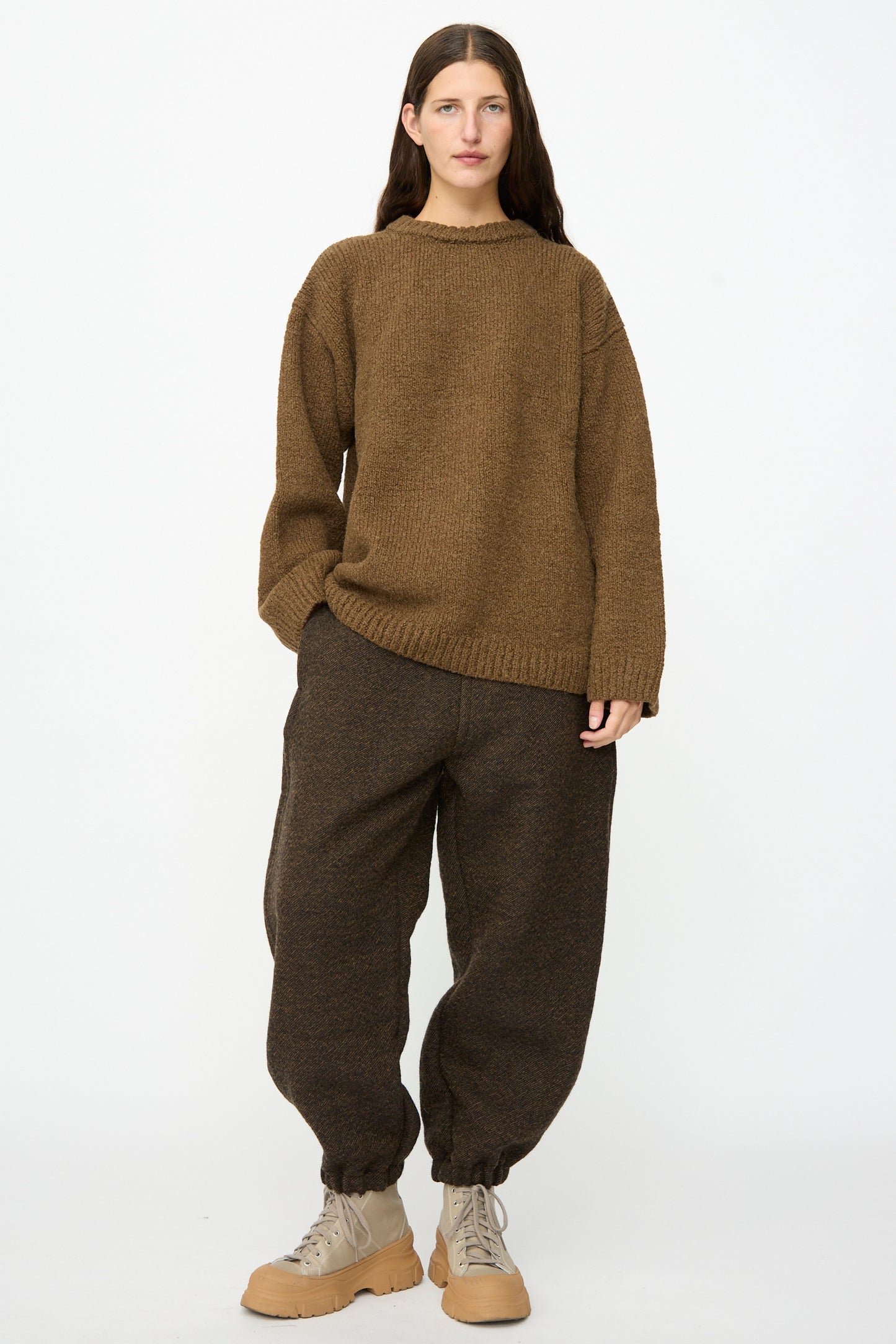 Person standing against a white background wearing a brown knit sweater, the Jamie Joggers in Blackmud from SONO with elastic cuffs, and beige lace-up boots.