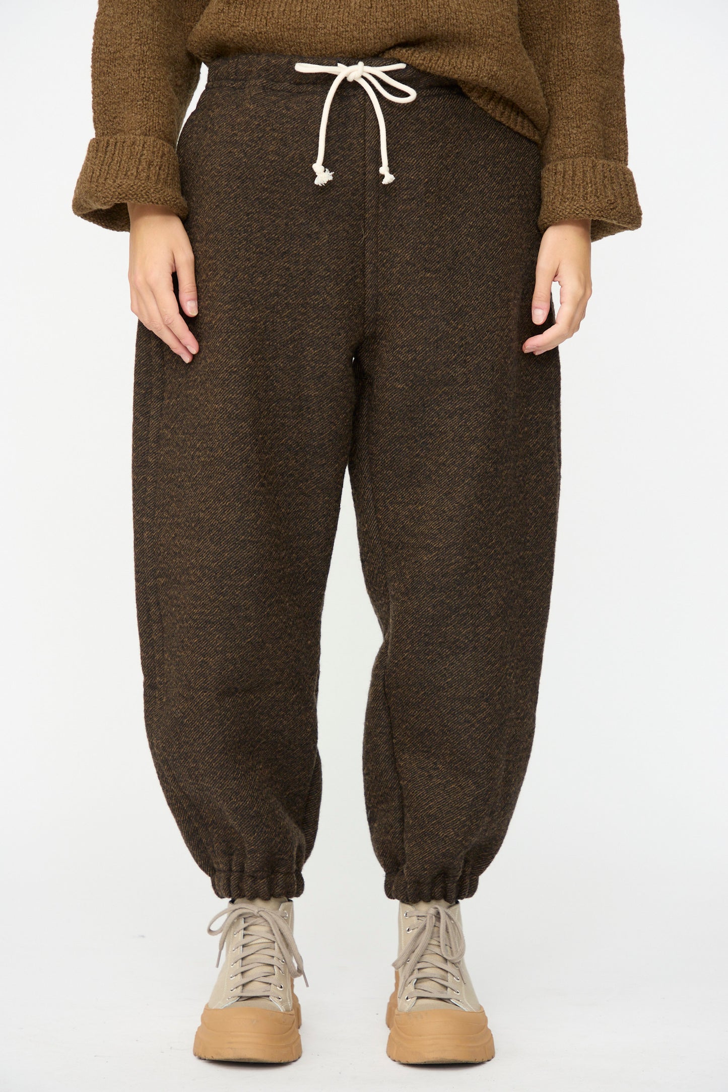 A person wearing the SONO Jamie Joggers in Blackmud, paired with a brown sweater and beige high-top sneakers, stands against a plain white background.