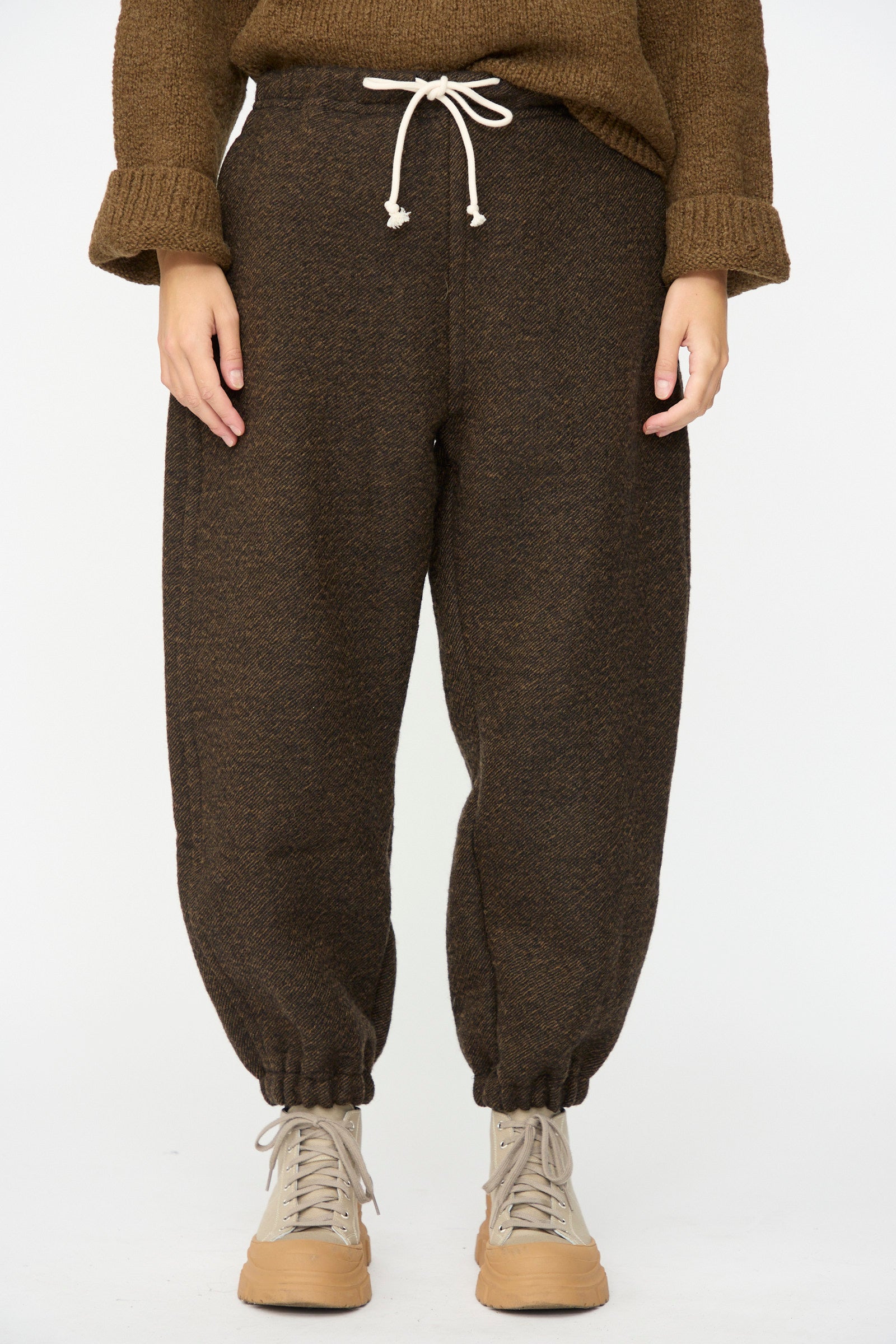 A person wearing the SONO Jamie Joggers in Blackmud, paired with a brown sweater and beige high-top sneakers, stands against a plain white background.