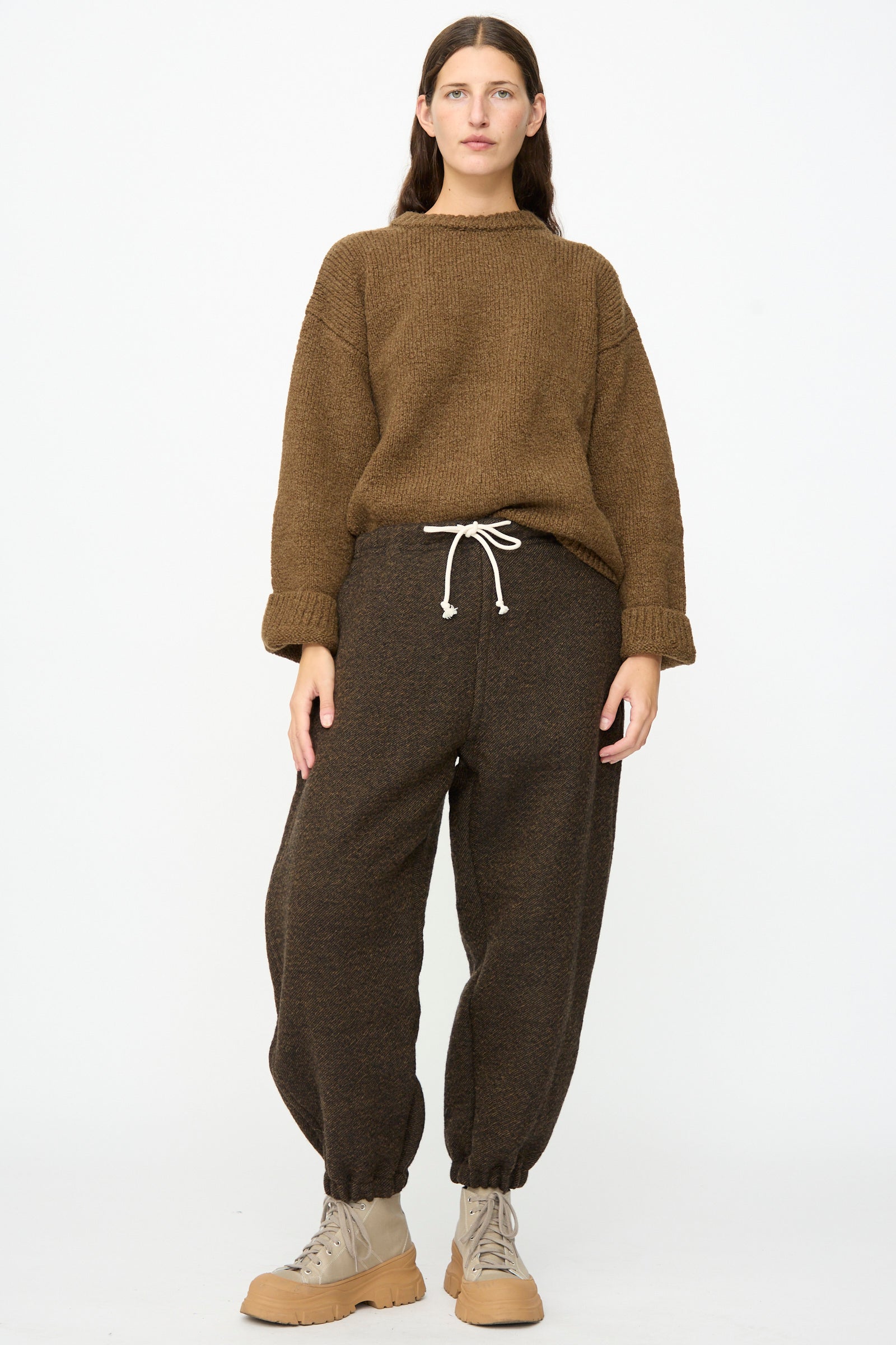 A person stands against a white background, wearing a brown knit sweater, SONO Jamie Joggers in Blackmud—loose, oversized jogger pants made in France—and beige lace-up boots.