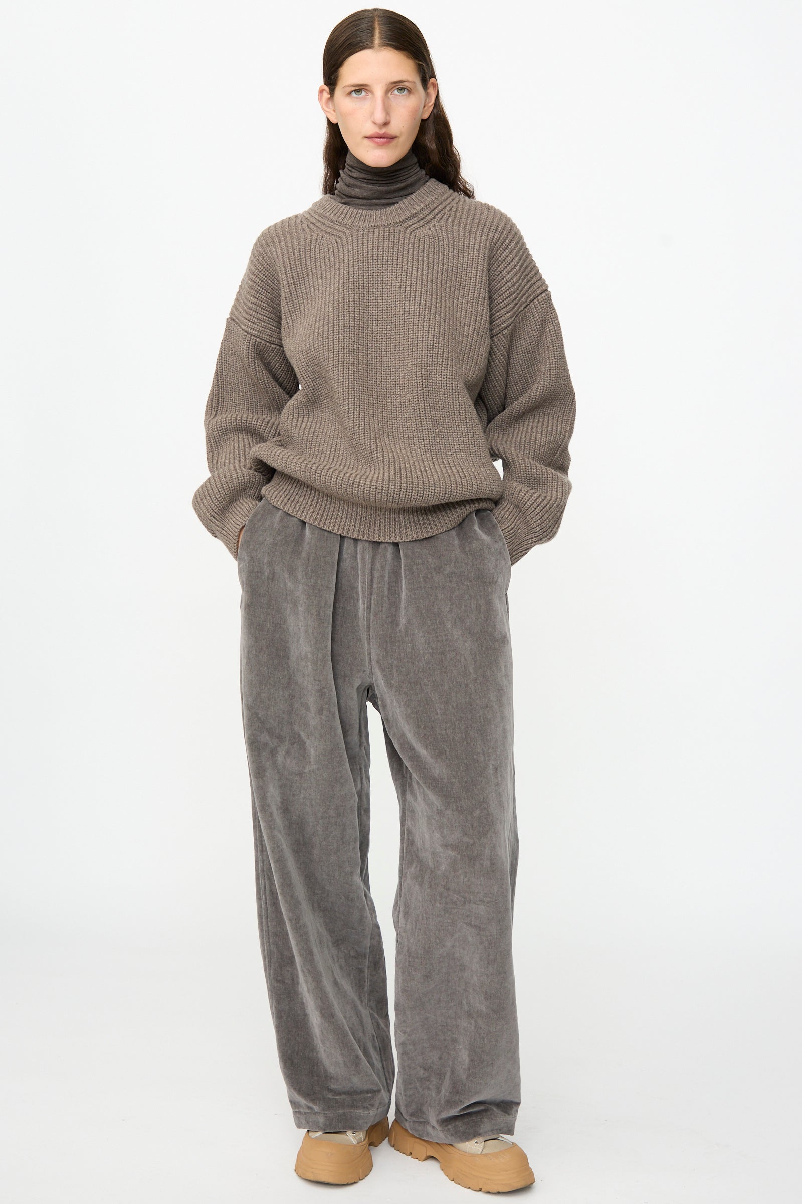 A person stands against a plain background with hands in pockets, wearing the SONO Joel Jumper in Smoke— a taupe ribbed knit wool crewneck sweater—paired with grey corduroy pants, a brown turtleneck underneath, and tan shoes.