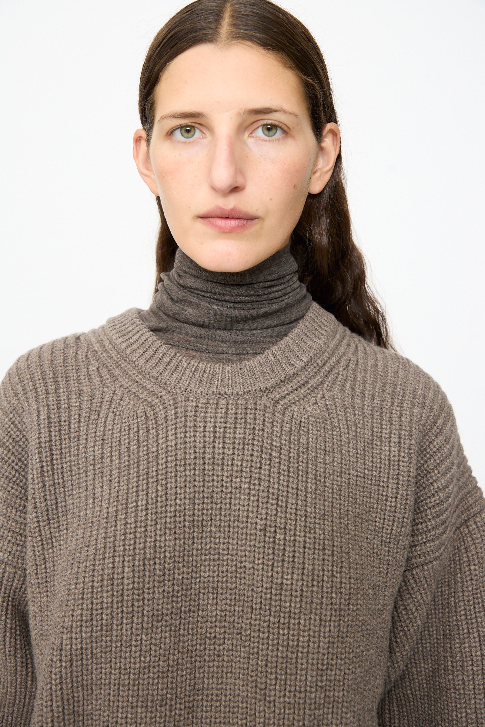 A person with long dark hair wears the SONO Joel Jumper in Smoke—a brown ribbed knit wool sweater—layered over a gray turtleneck, standing facing the camera against a plain light background.