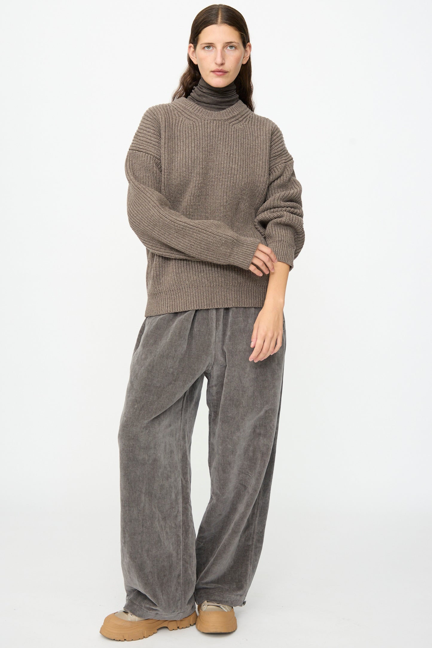 A person stands against a plain background, wearing the SONO Joel Jumper in Smoke—an oversized ribbed knit wool pullover—layered over a dark turtleneck, with loose gray pants and tan shoes.