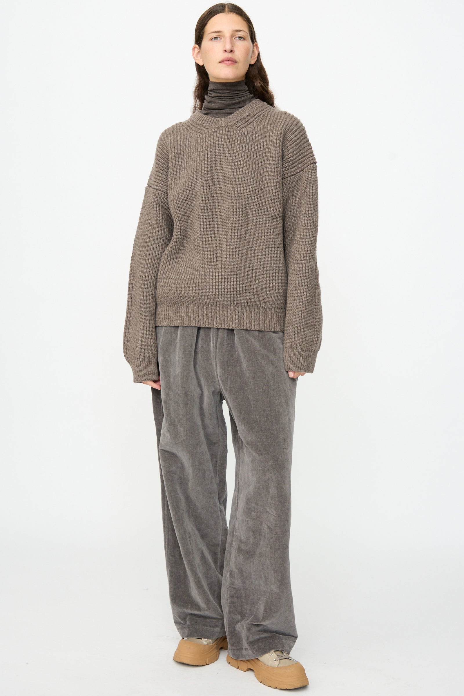 A person with long, center-parted hair stands against a plain background, wearing the Joel Jumper in Smoke by SONO layered over a dark turtleneck, paired with wide gray corduroy pants and tan shoes.