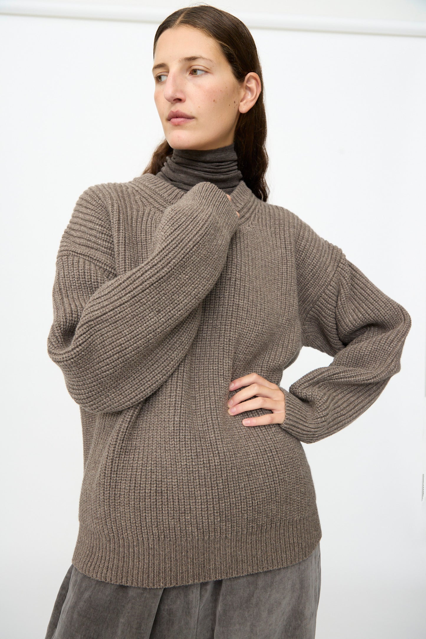 A person wearing the SONO Joel Jumper in Smoke and gray pants stands against a plain white background, looking to the side with one hand on their hip.