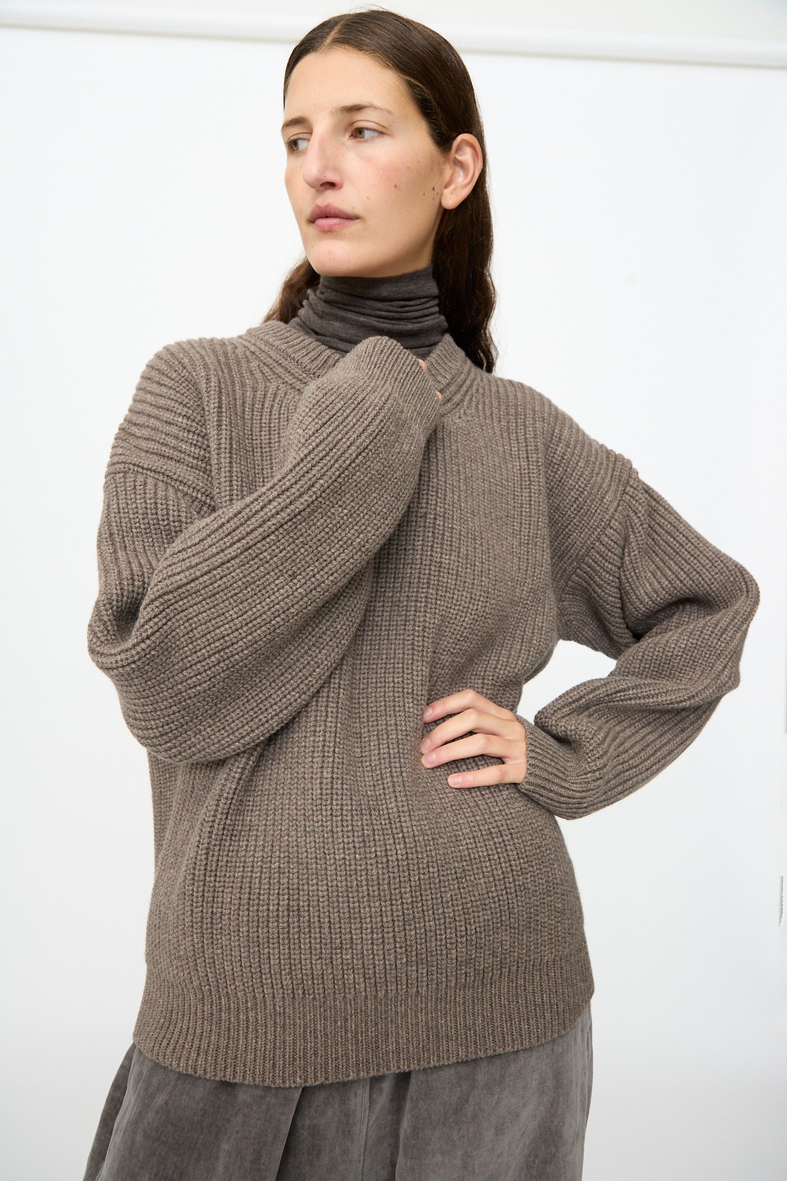 A person wearing the SONO Joel Jumper in Smoke and gray pants stands against a plain white background, looking to the side with one hand on their hip.