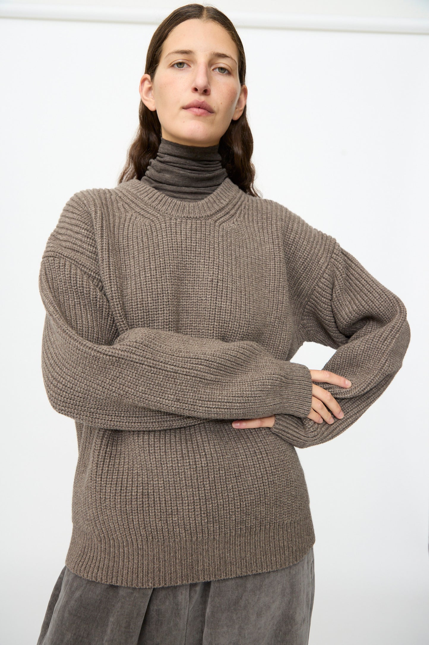A person with long brown hair wears the SONO Joel Jumper in Smoke over a brown turtleneck and a gray skirt, standing against a plain white background.