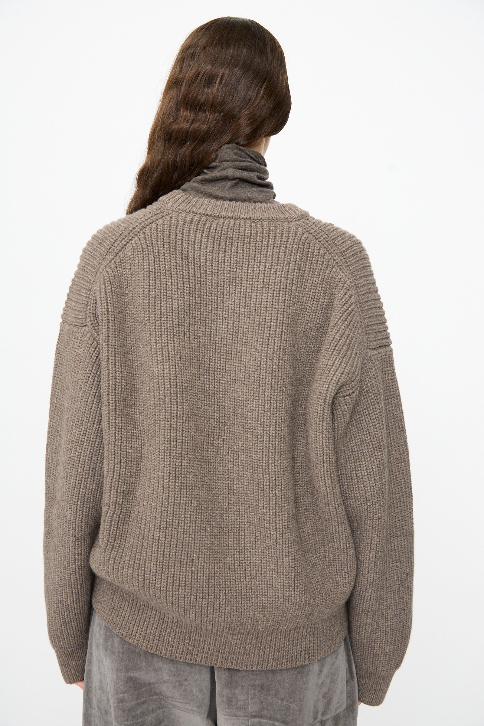 A person with long brown hair stands facing away against a plain white background, wearing the Joel Jumper in Smoke by SONO—a thick, ribbed knit wool taupe sweater—layered over a gray turtleneck top and paired with gray velvety pants.