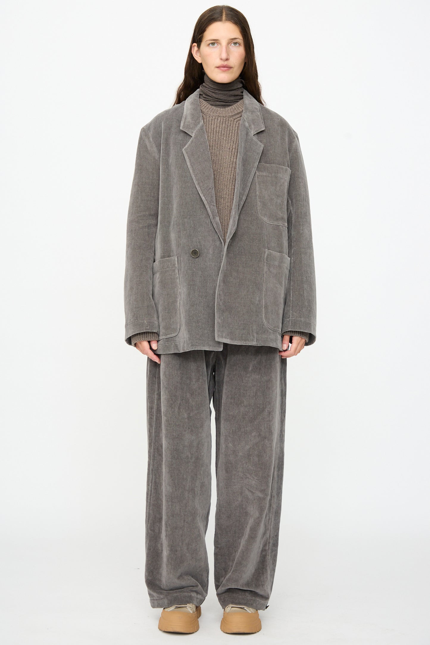 A person stands against a plain white background wearing an oversized gray corduroy suit, brown sweater, and tan shoes, with the look completed by the SONO Juri Jacket in Elephant—a statement oversized jacket crafted from washed velvet.