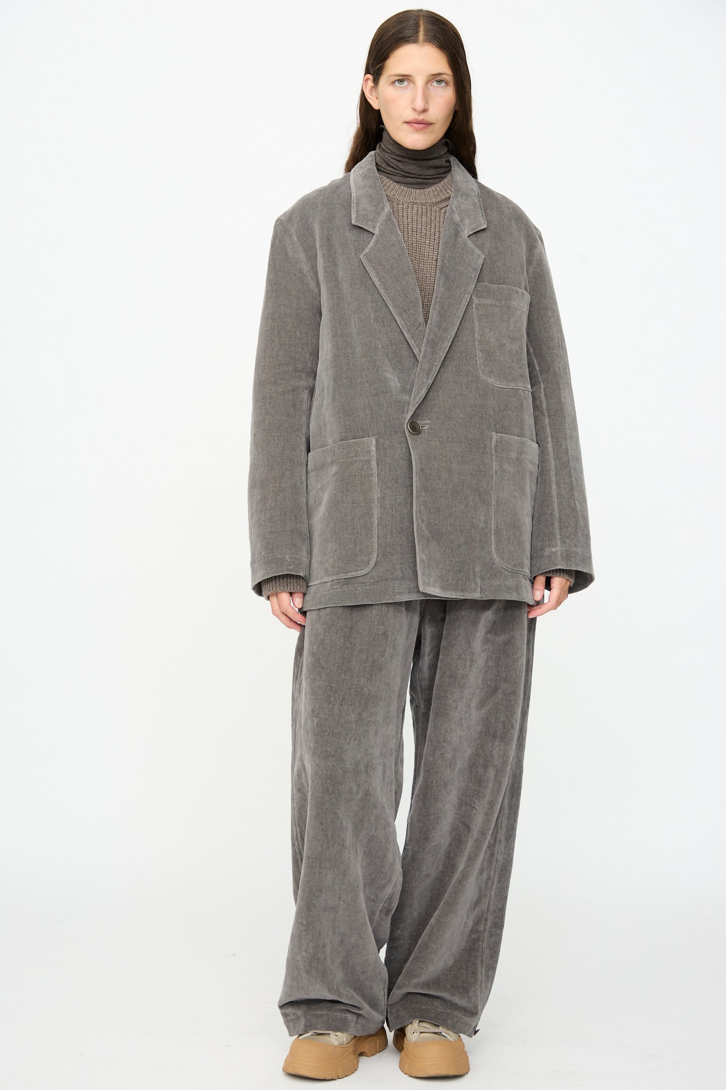 A person standing against a white background is wearing the SONO Juri Jacket in Elephant, an oversized grey corduroy jacket, paired with a brown organic cotton turtleneck sweater and tan shoes.
