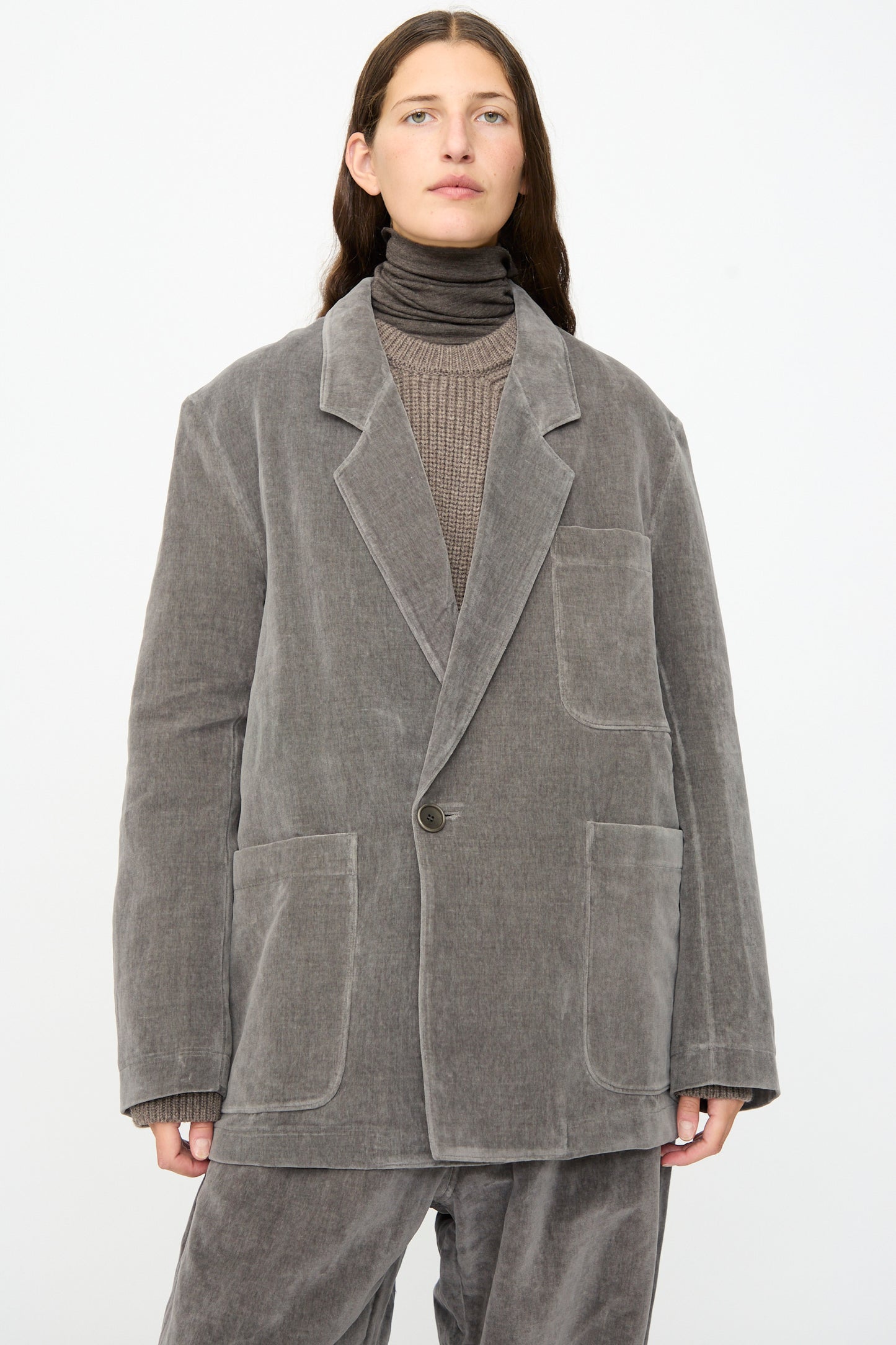 A person wears the SONO Juri Jacket in Elephant—an oversized gray corduroy blazer crafted from organic cotton—paired with matching pants, a taupe ribbed sweater, and a dark turtleneck against a plain white background.