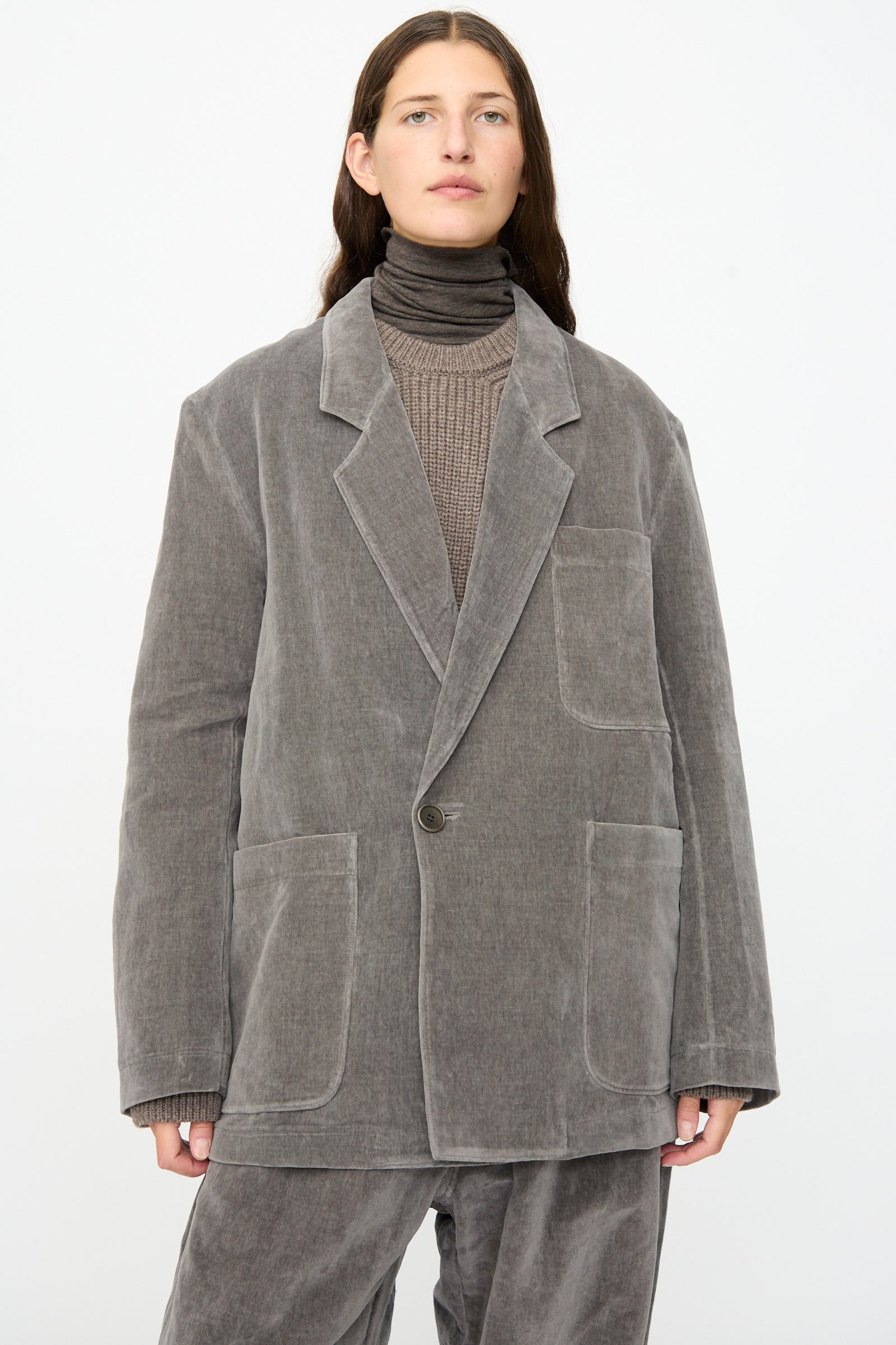 A person wears the SONO Juri Jacket in Elephant—an oversized gray corduroy blazer crafted from organic cotton—paired with matching pants, a taupe ribbed sweater, and a dark turtleneck against a plain white background.
