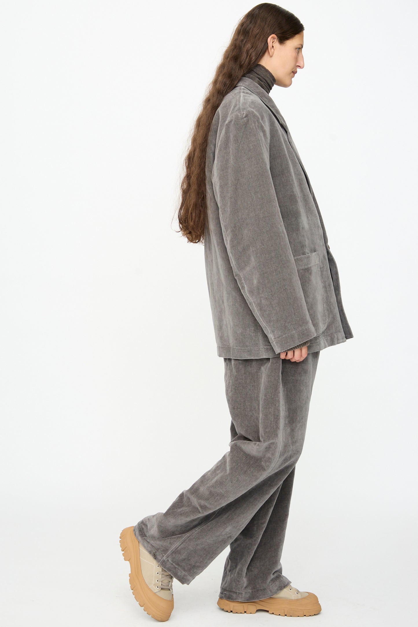 A person with long brown hair is wearing the SONO Juri Jacket in Elephant—a loose-fitting grey corduroy suit and oversized jacket—paired with a brown turtleneck and beige chunky shoes, walking against a plain white background.
