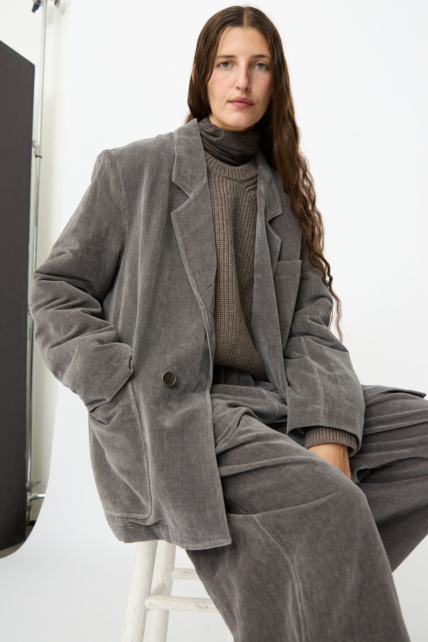 A woman with long brown hair wears the SONO Juri Jacket in Elephant, an oversized gray corduroy jacket crafted from organic cotton, styled over a taupe turtleneck sweater and coordinating suit. She is seated on a white stool against a plain studio background.