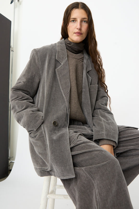 A woman with long brown hair wears the SONO Juri Jacket in Elephant, an oversized gray corduroy jacket crafted from organic cotton, styled over a taupe turtleneck sweater and coordinating suit. She is seated on a white stool against a plain studio background.