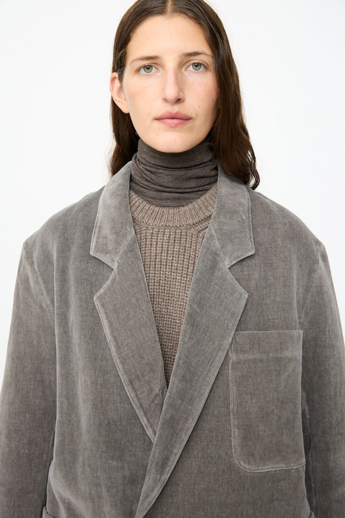 A person wearing the SONO Juri Jacket in Elephant—an oversized grey jacket crafted from organic cotton—is layered over a brown turtleneck and knit sweater, standing against a plain white background.