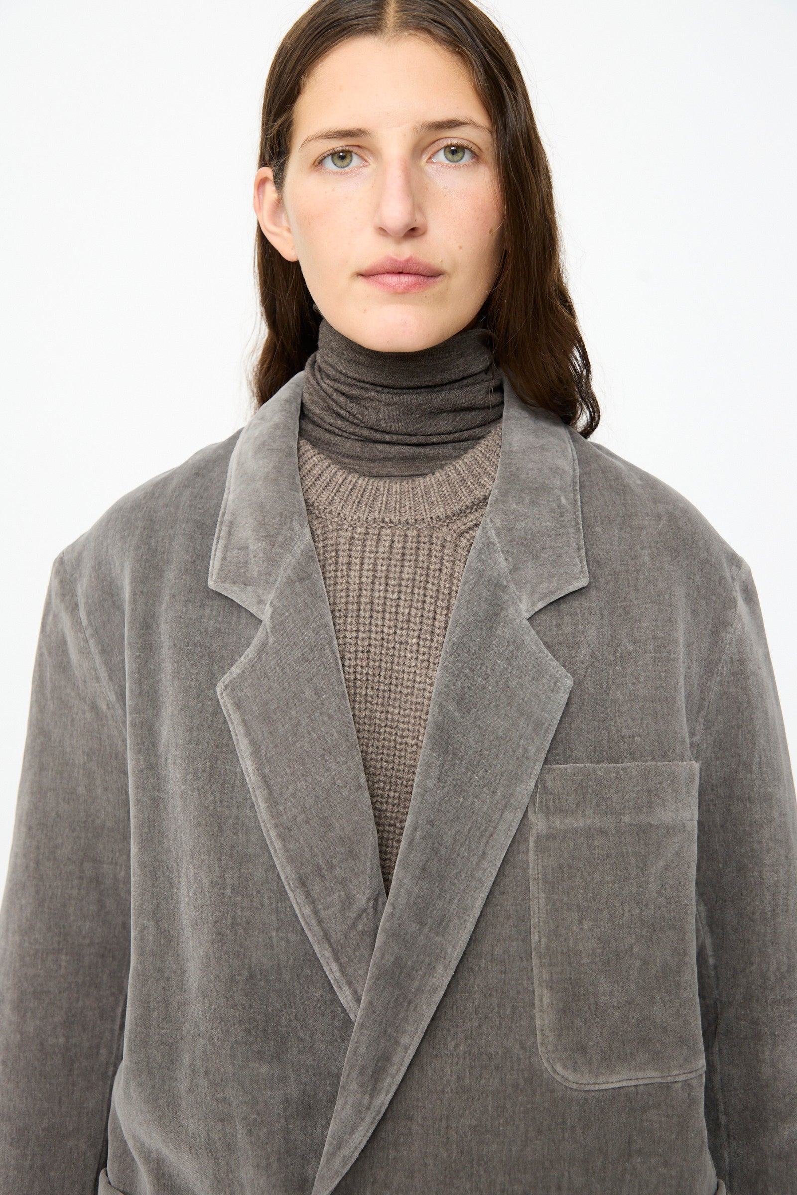 A person wearing the SONO Juri Jacket in Elephant—an oversized grey jacket crafted from organic cotton—is layered over a brown turtleneck and knit sweater, standing against a plain white background.