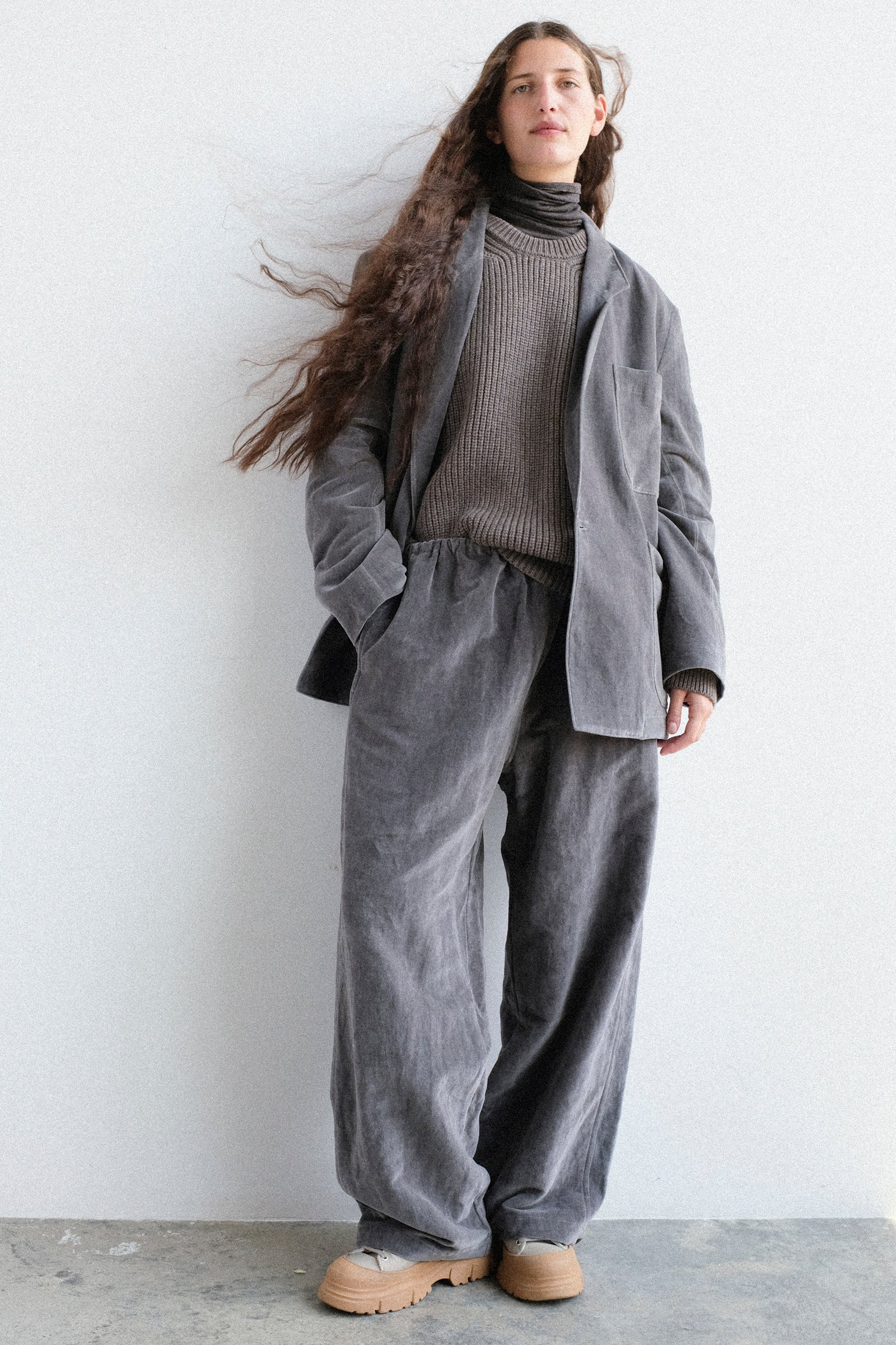 A woman stands against a plain light gray wall wearing SONO's Juri Jacket, Joel Jumper, and Sol Snowpant.