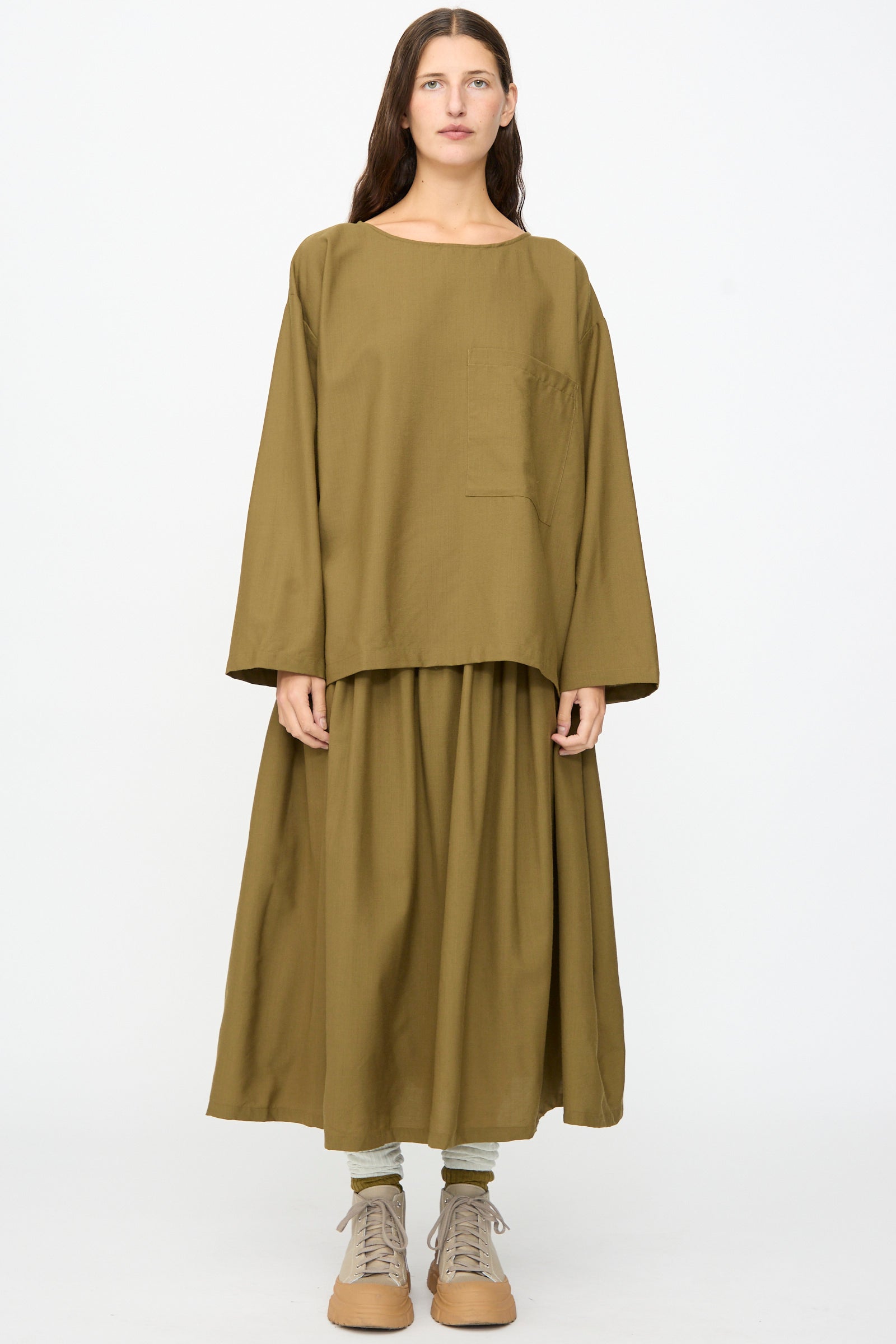 A person stands facing forward against a plain white background, wearing the SONO Laik Longsleeve in Kelp—an oversized fit olive brown washed wool shirt featuring a chest pocket—paired with a matching wide skirt, green socks, and tan boots. 