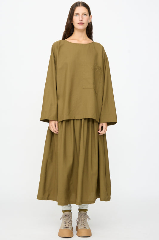 A person stands facing forward against a plain white background, wearing the SONO Laik Longsleeve in Kelp—an oversized fit olive brown washed wool shirt featuring a chest pocket—paired with a matching wide skirt, green socks, and tan boots. 