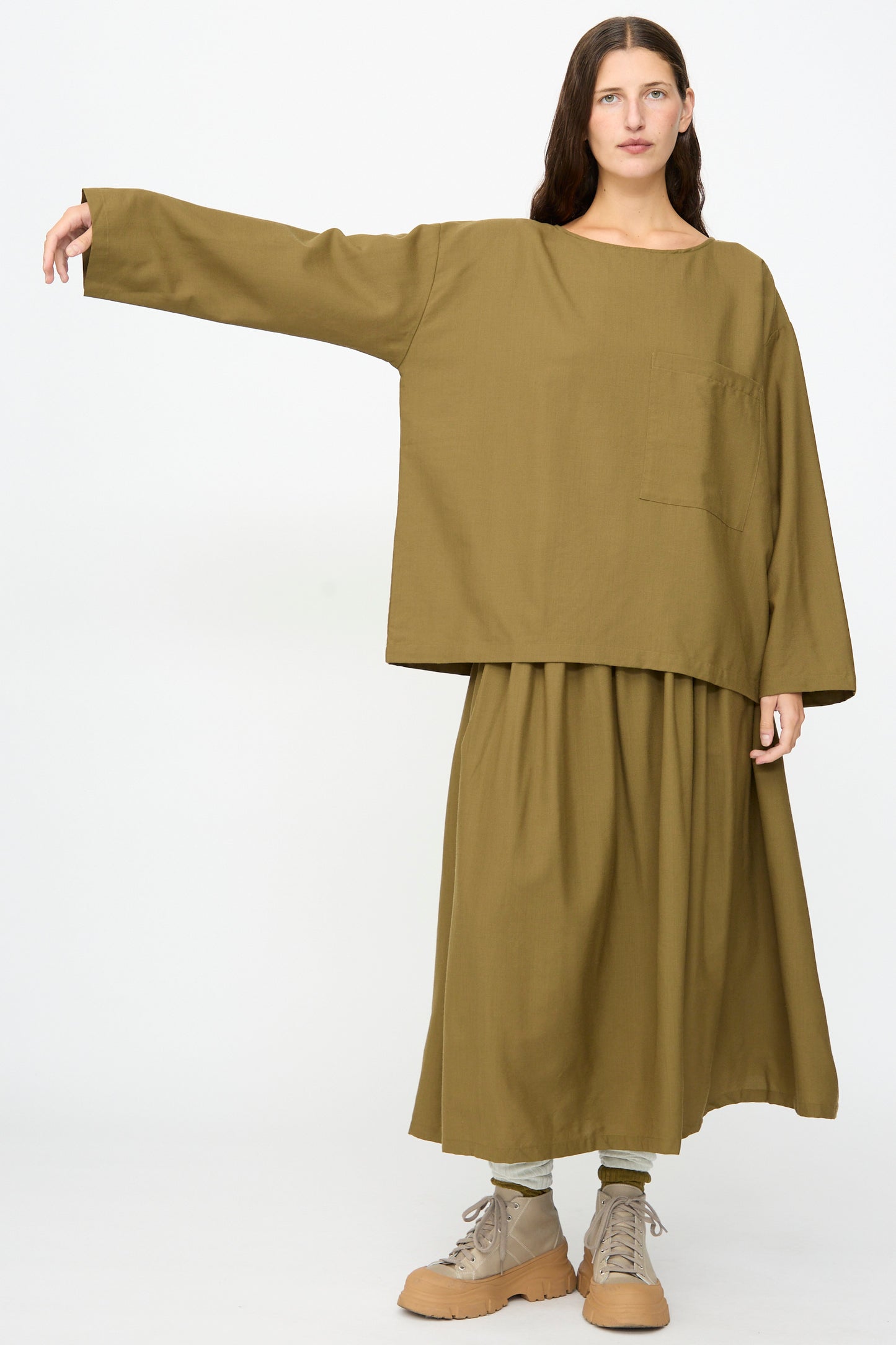 A woman stands against a plain background wearing the SONO Laik Longsleeve in Kelp—an oversized olive green top featuring a pocket—paired with a matching skirt and tan lace-up shoes, her left arm extended sideways.