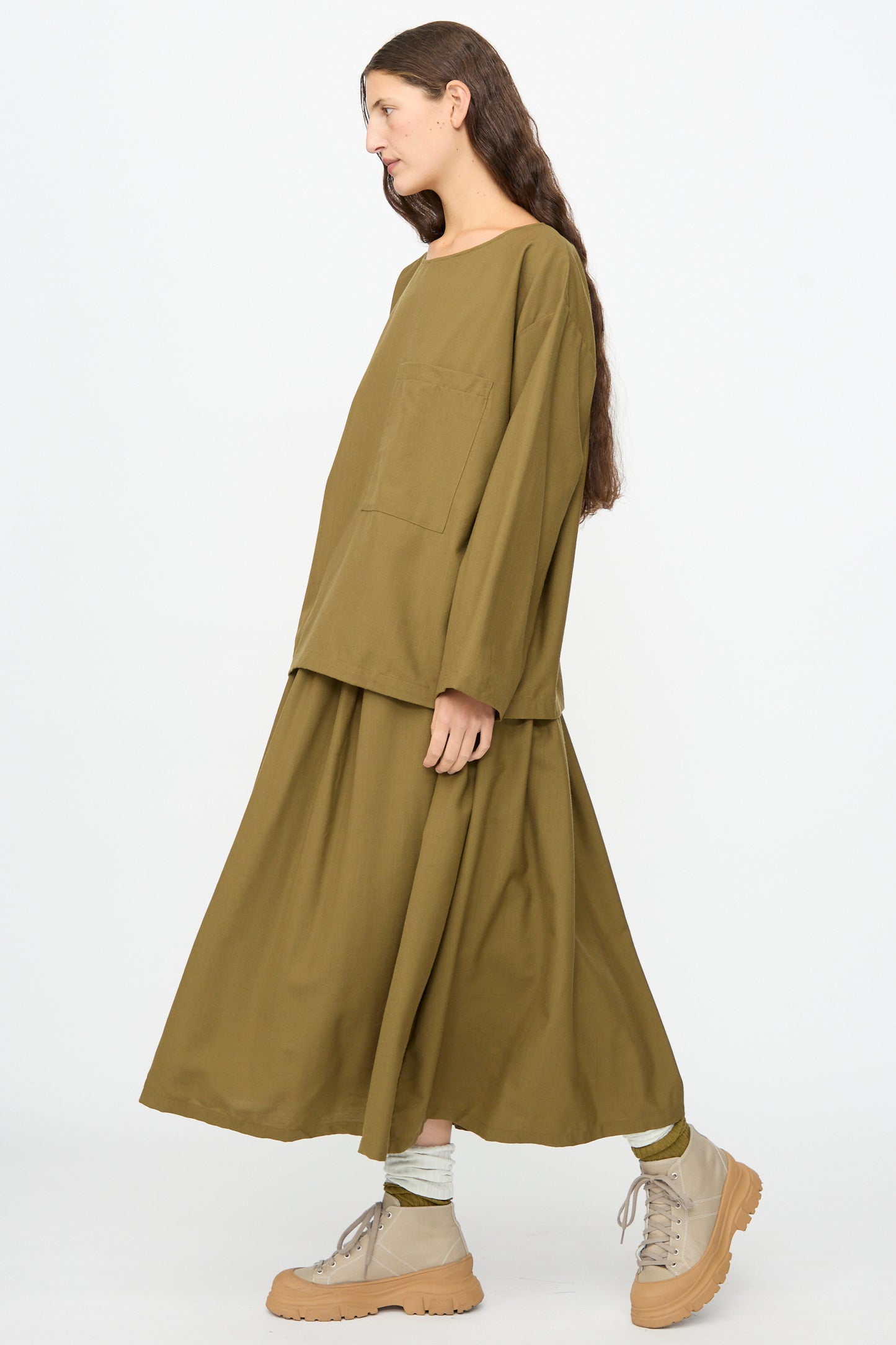 A person with long brown hair wears the SONO Laik Longsleeve in Kelp, styled in an oversized fit, along with a matching maxi skirt, beige socks, and tan chunky shoes, standing against a plain white background.