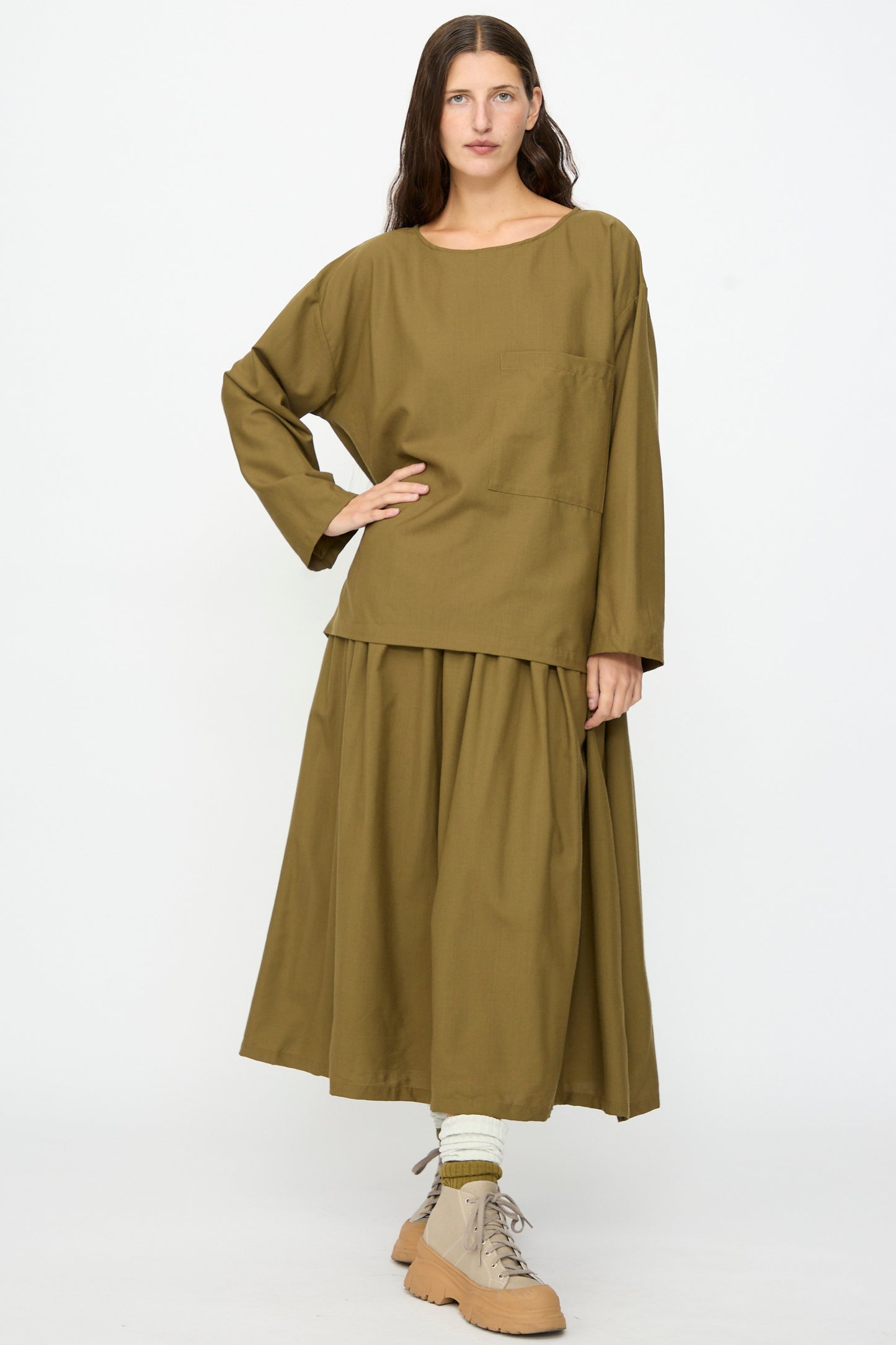 Wearing the SONO Laik Longsleeve in Kelp, styled with a coordinating ankle-length skirt, beige socks, and tan lace-up shoes, a person stands against a plain white background.