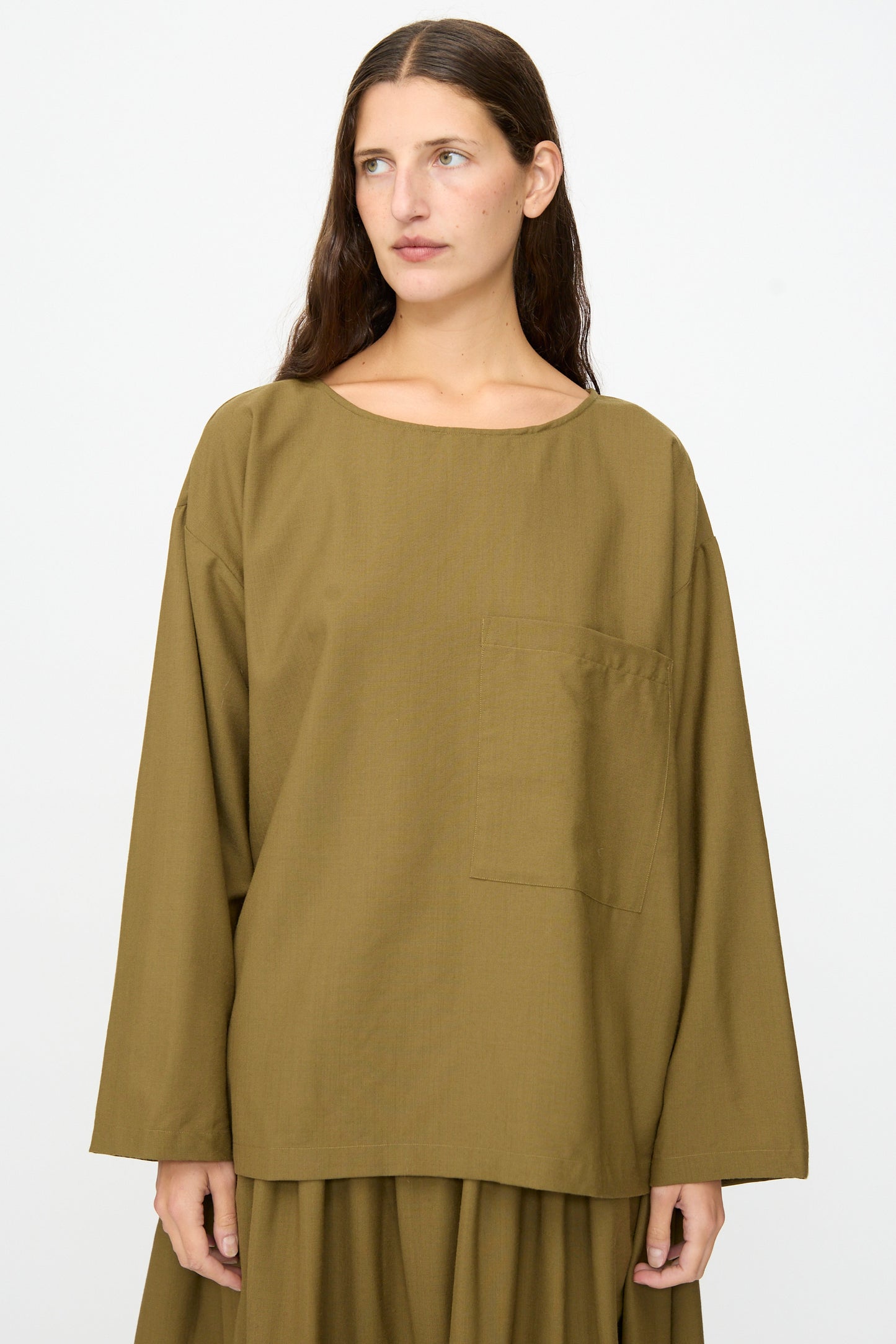 A person with long brown hair wears the SONO Laik Longsleeve in Kelp, a washed wool shirt with a front pocket, styled with a matching oversized skirt, standing against a plain white background.