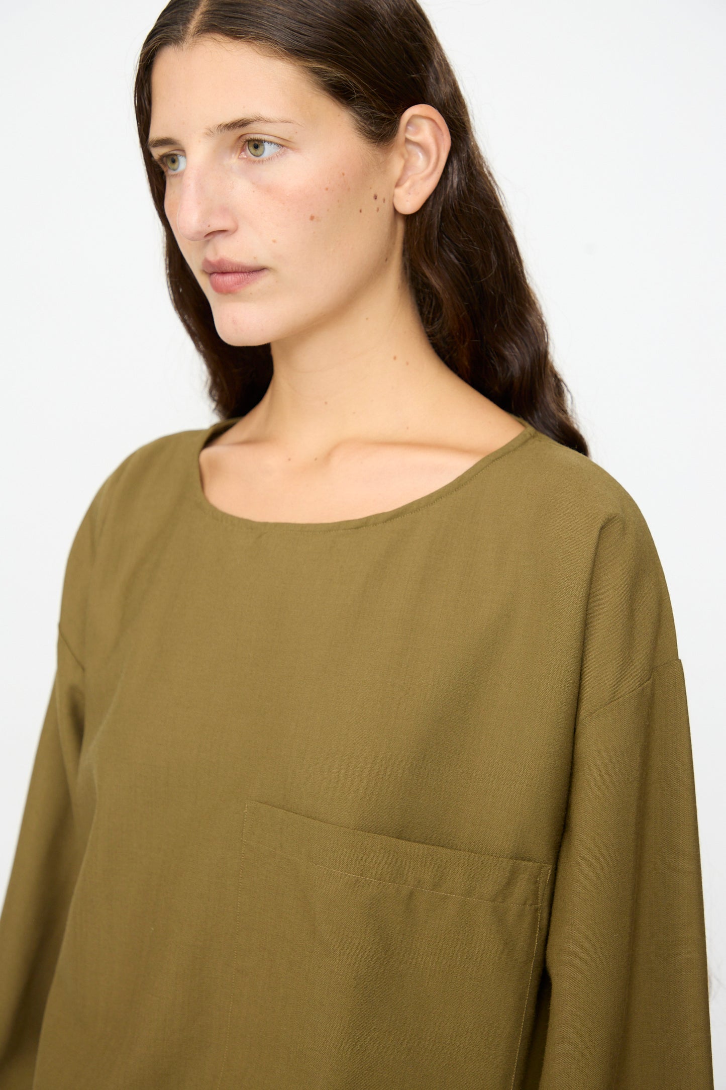 A woman with long brown hair wears the SONO Laik Longsleeve in Kelp, an olive green, oversized washed wool shirt featuring long sleeves and a chest pocket. She looks slightly to the left against a plain background. This piece is made in France.