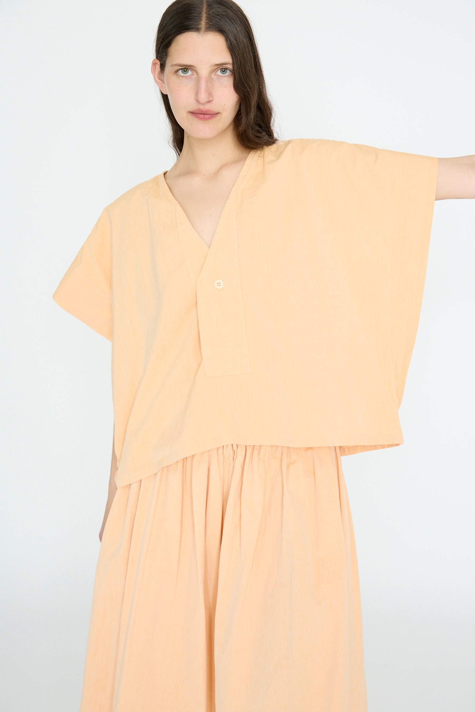 A person stands against a plain background wearing the SONO Louise Shirt in Dusty Peach, a loose-fitting, organic cotton top with corozo nut buttons and short sleeves, paired with a matching skirt.