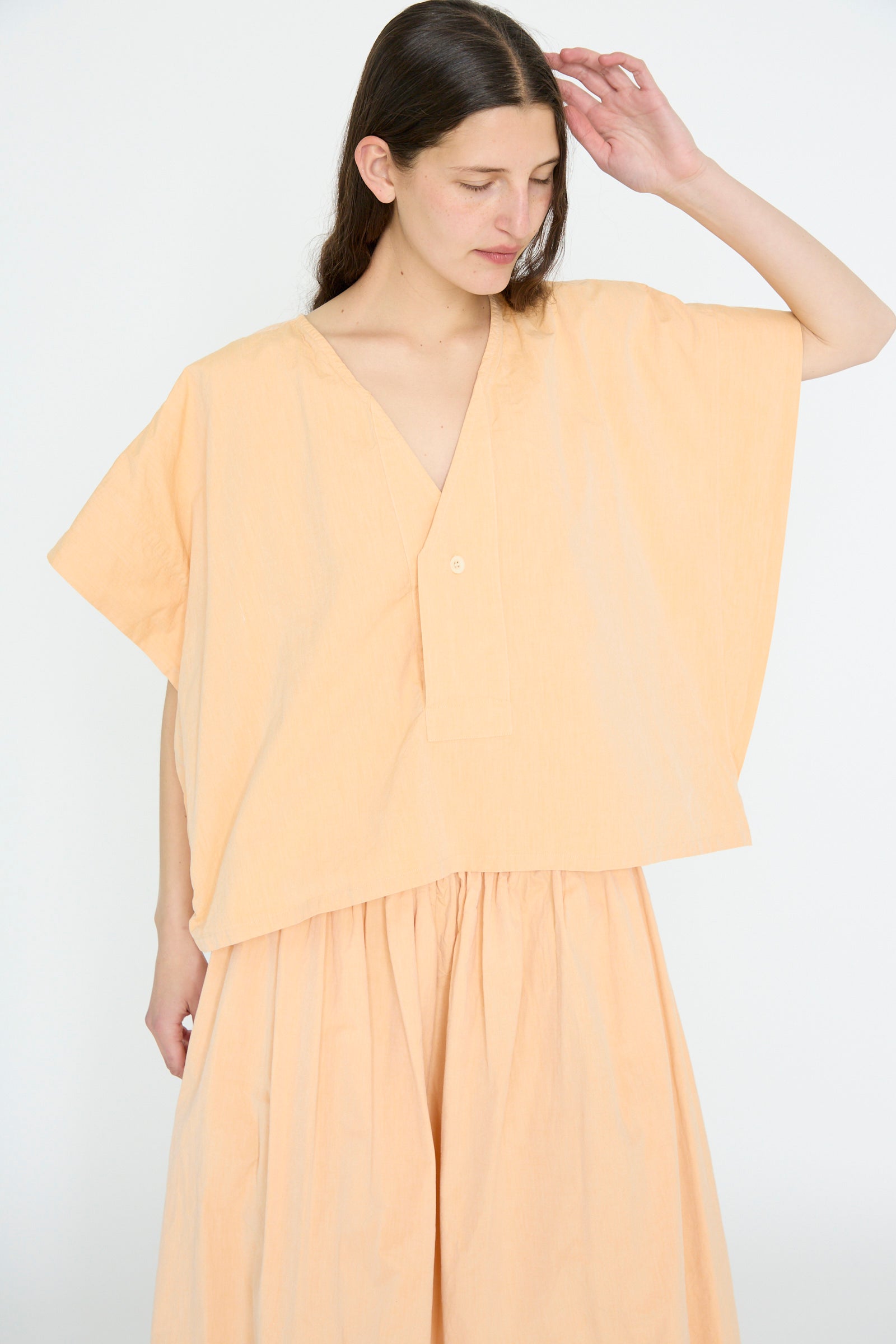 A woman with long brown hair wears the SONO Louise Shirt in Dusty Peach—an organic cotton, loose-fitting cropped placket shirt with corozo nut buttons—and a matching skirt, standing against a plain white background.