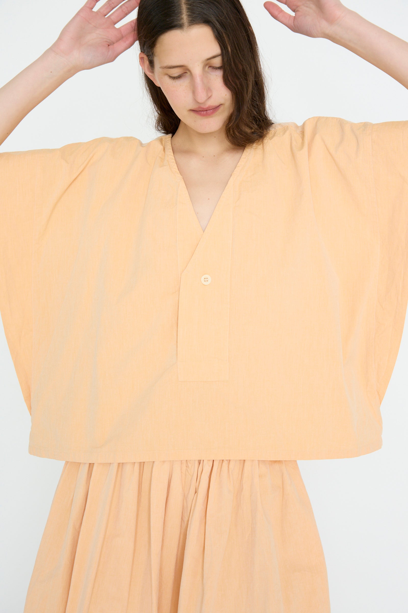 A person wearing the SONO Louise Shirt in Dusty Peach and an organic cotton skirt stands against a plain background, eyes closed and hands raised near their head.