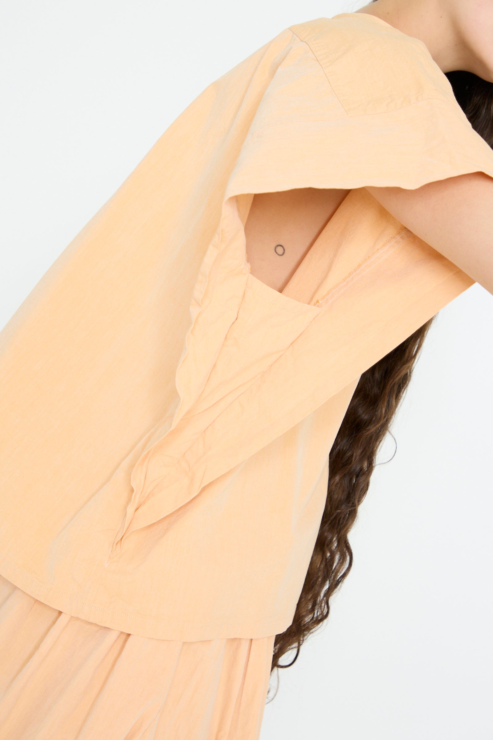 Wearing the SONO Louise Shirt in Dusty Peach, a loose cropped shirt with corozo nut buttons and an open back, revealing a small side tattoo and part of long hair.