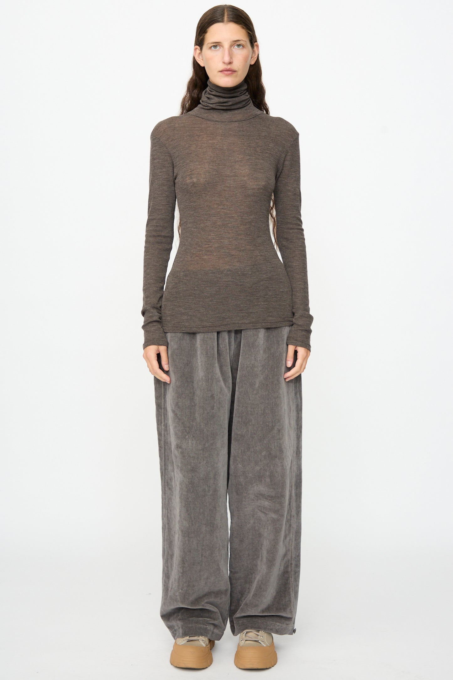 A person stands facing forward, wearing the SONO Roberta Rollneck in Charcoal, loose gray velvet pants, and beige shoes against a plain white background.
