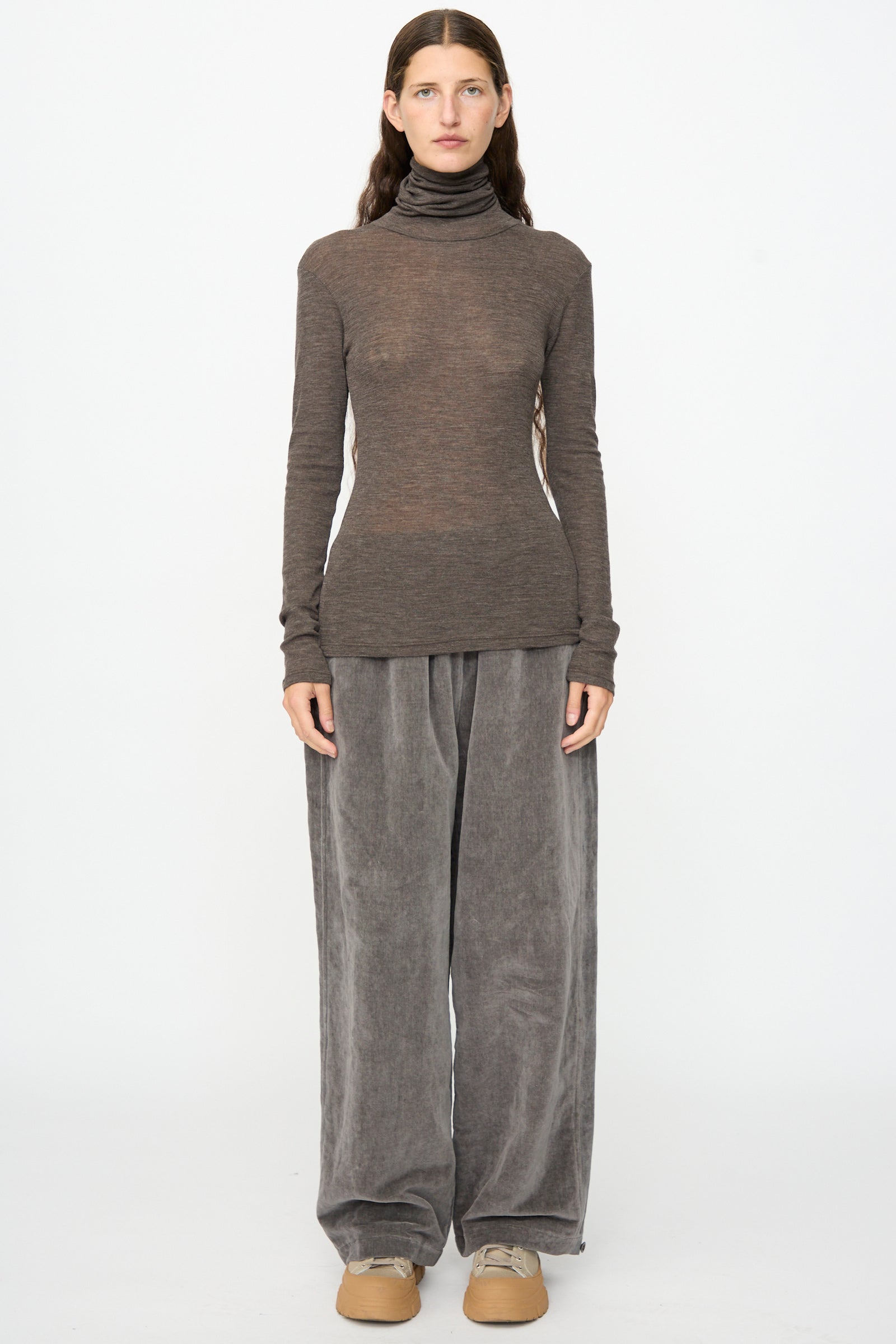 A person stands facing forward, wearing the SONO Roberta Rollneck in Charcoal, loose gray velvet pants, and beige shoes against a plain white background.
