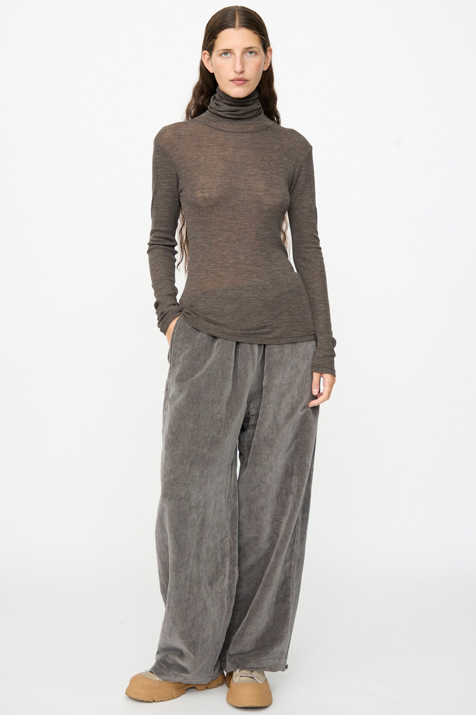 A person stands against a plain background, wearing the SONO Roberta Rollneck in Charcoal—a slim fit rolled turtleneck—paired with loose gray pants and beige shoes, hands in pockets and long hair down.
