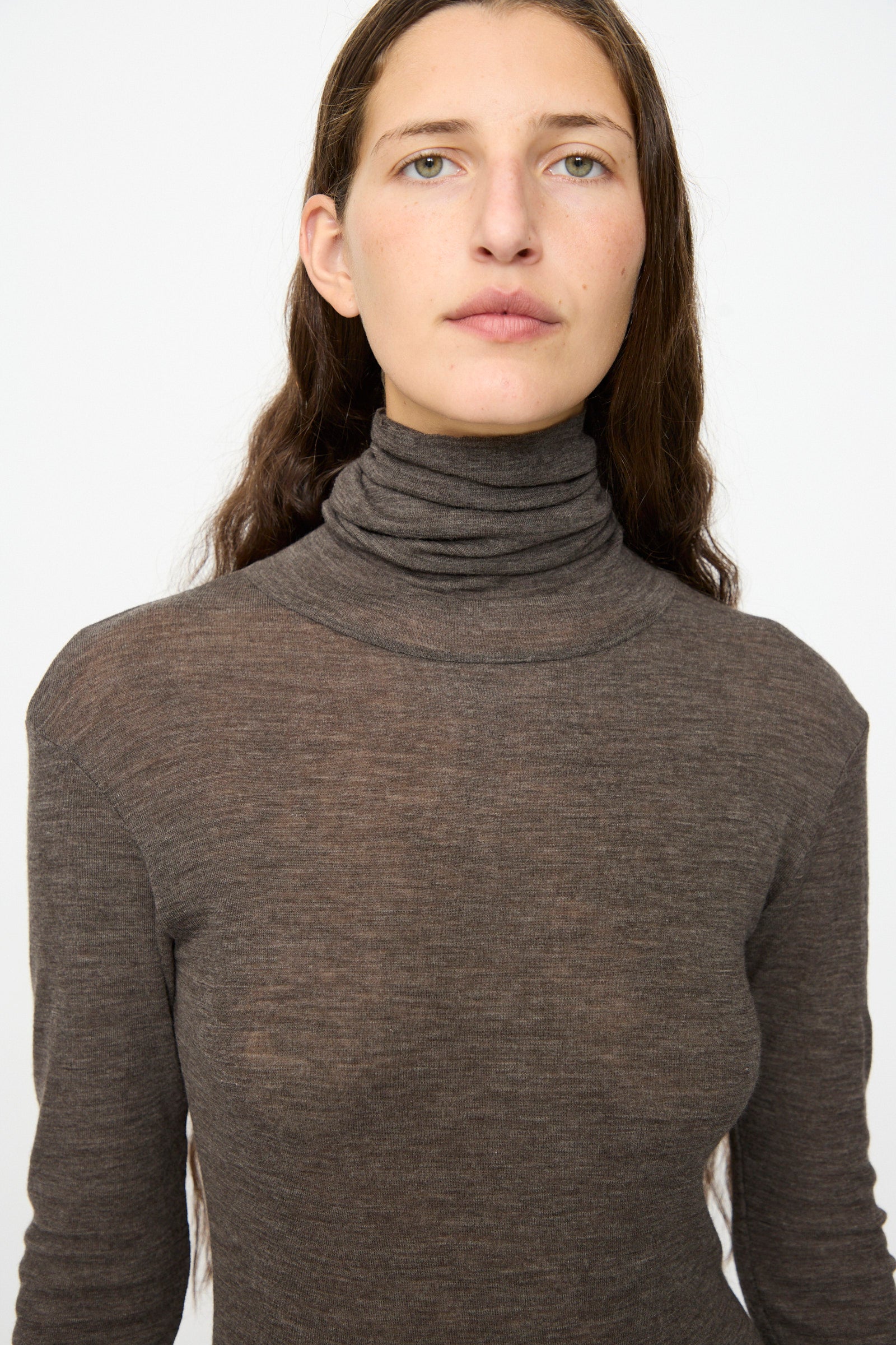 A person with long brown hair wears the SONO Roberta Rollneck in Charcoal, a slim fit sheer turtleneck, and looks directly at the camera against a plain white background.