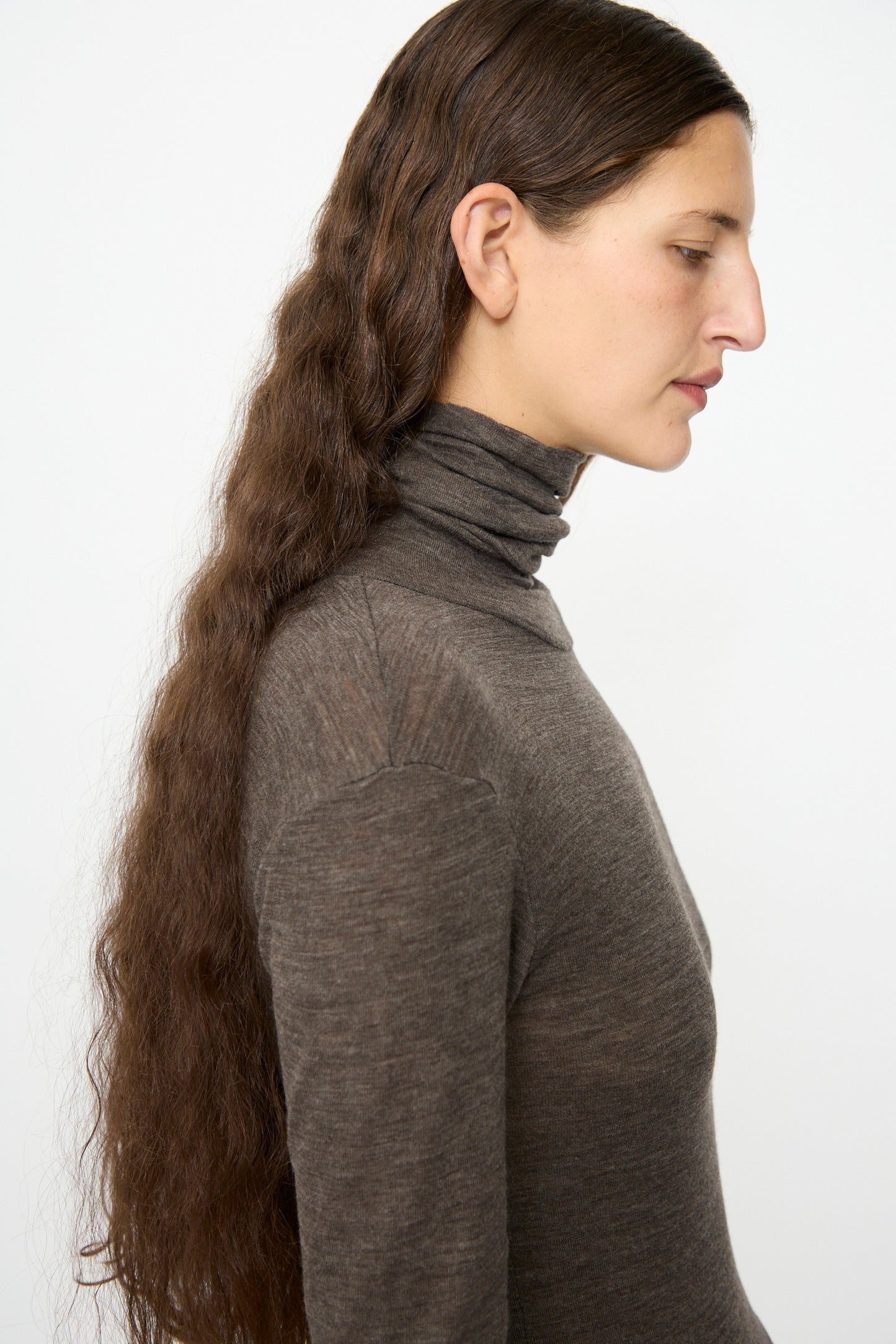 A person with long, wavy brown hair stands in profile against a plain white background, wearing the SONO Roberta Rollneck in Charcoal—a slim fit merino wool shirt with a rolled turtleneck.