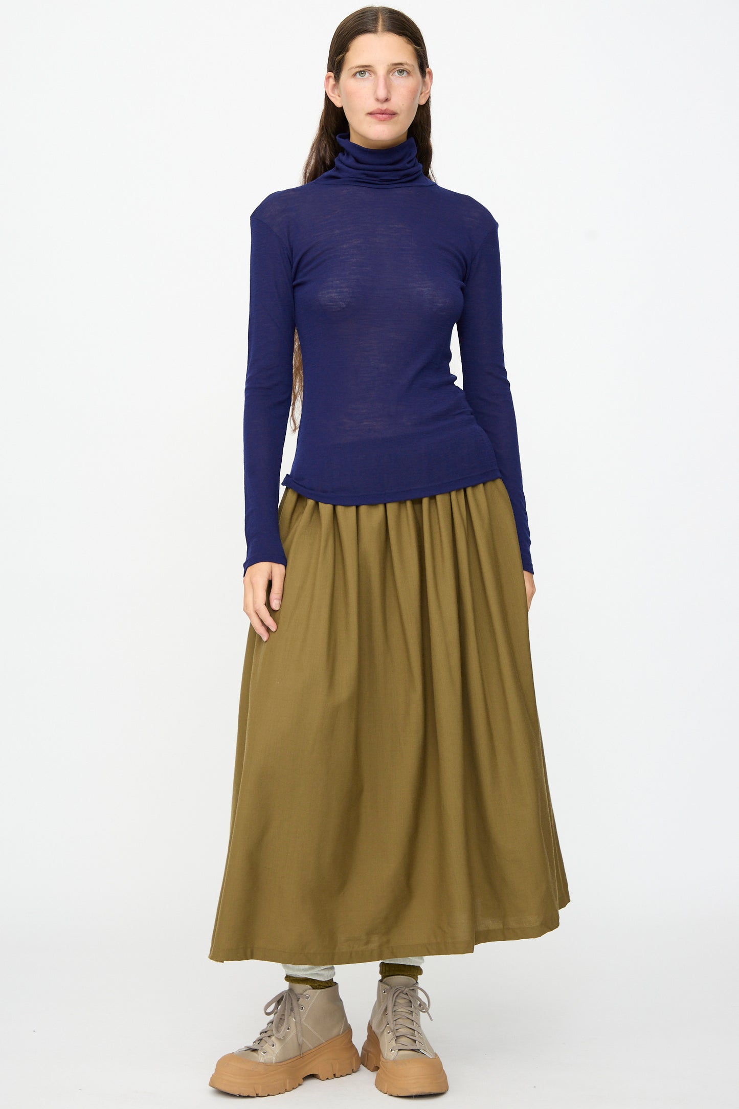 A person stands facing forward against a plain white background, wearing the SONO Roberta Rollneck in Indigo paired with an olive green long skirt and tan lace-up shoes.