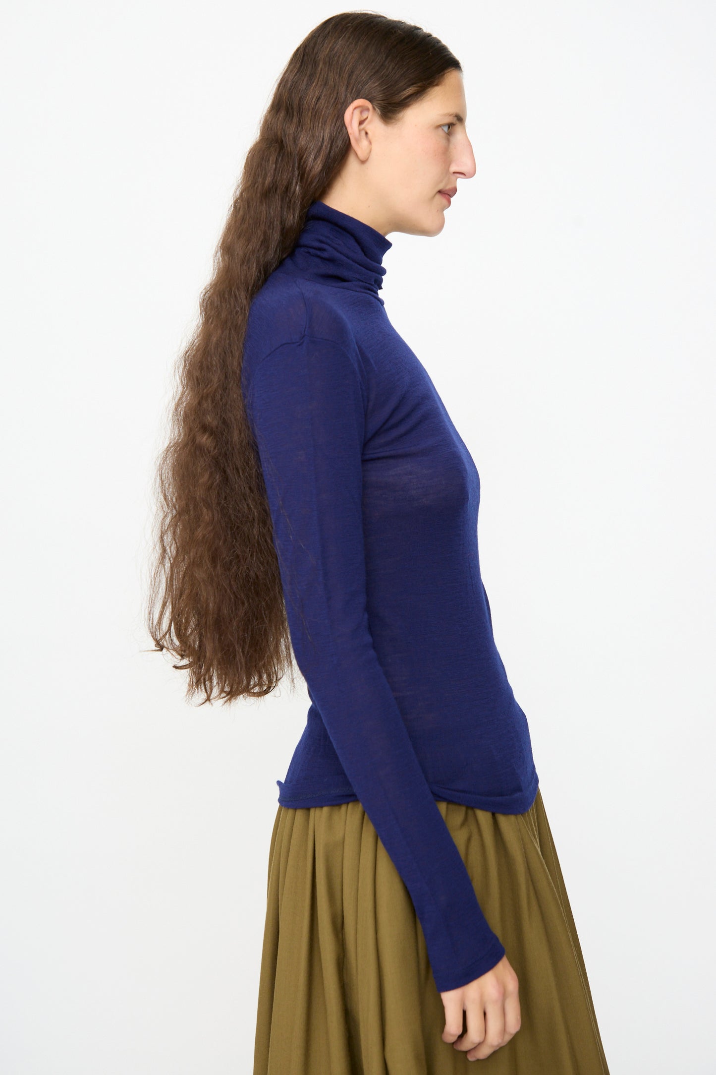 A person with long brown hair, wearing the SONO Roberta Rollneck in Indigo and an olive-green skirt, stands in profile against a plain white background.