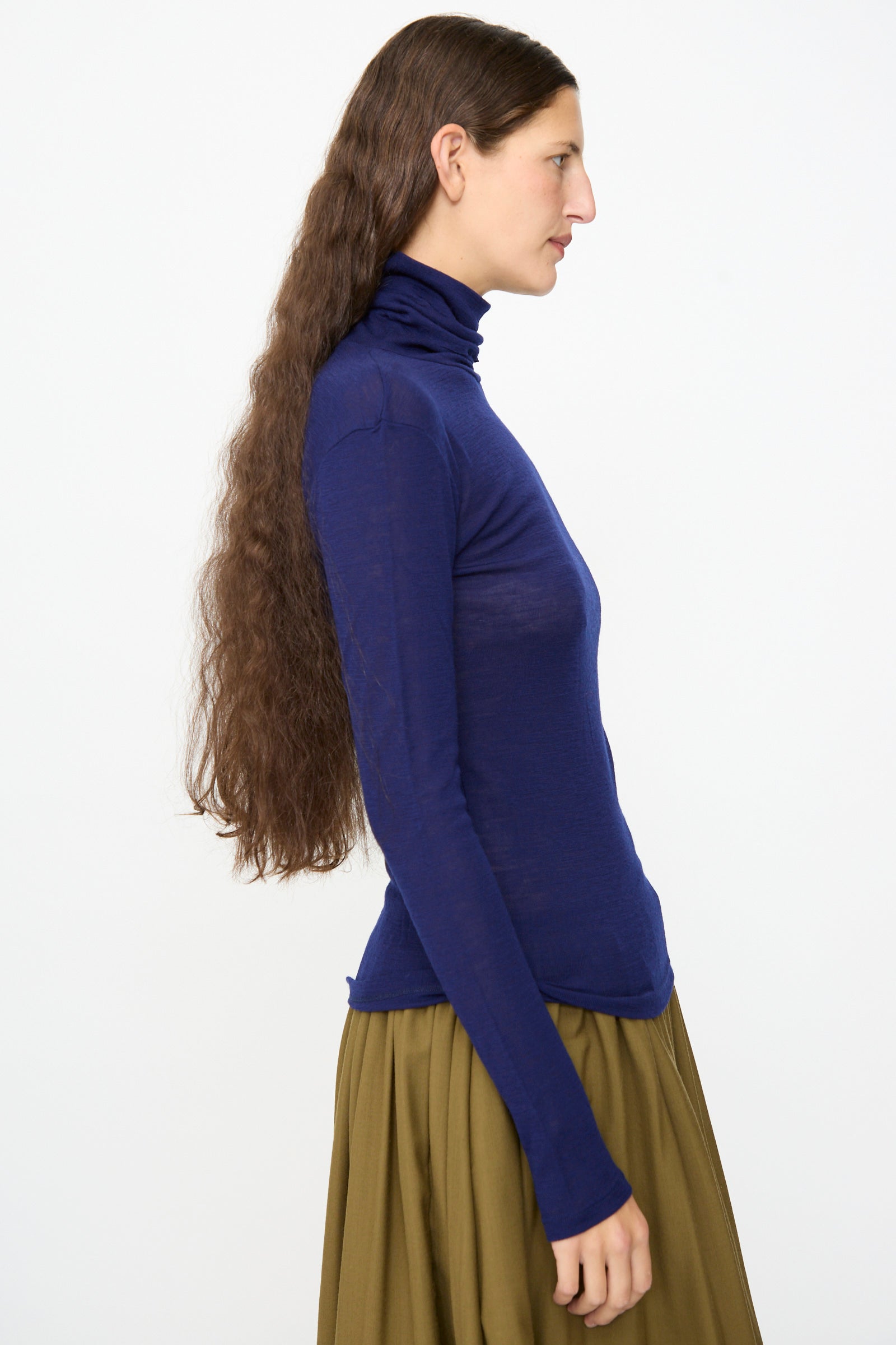 A person with long brown hair, wearing the SONO Roberta Rollneck in Indigo and an olive-green skirt, stands in profile against a plain white background.
