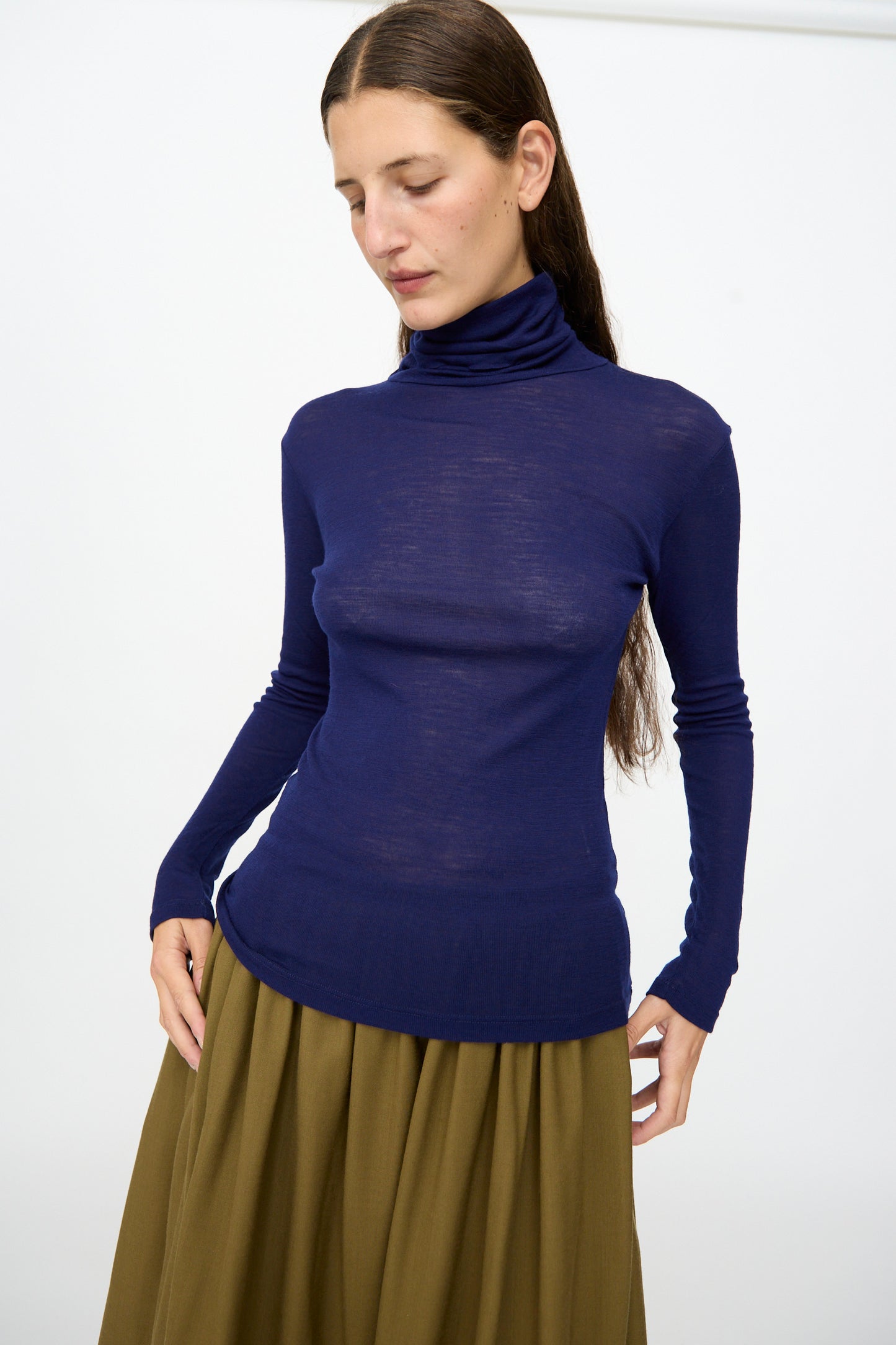 A woman with long brown hair wears the SONO Roberta Rollneck in Indigo, paired with an olive green skirt, standing against a plain white background.